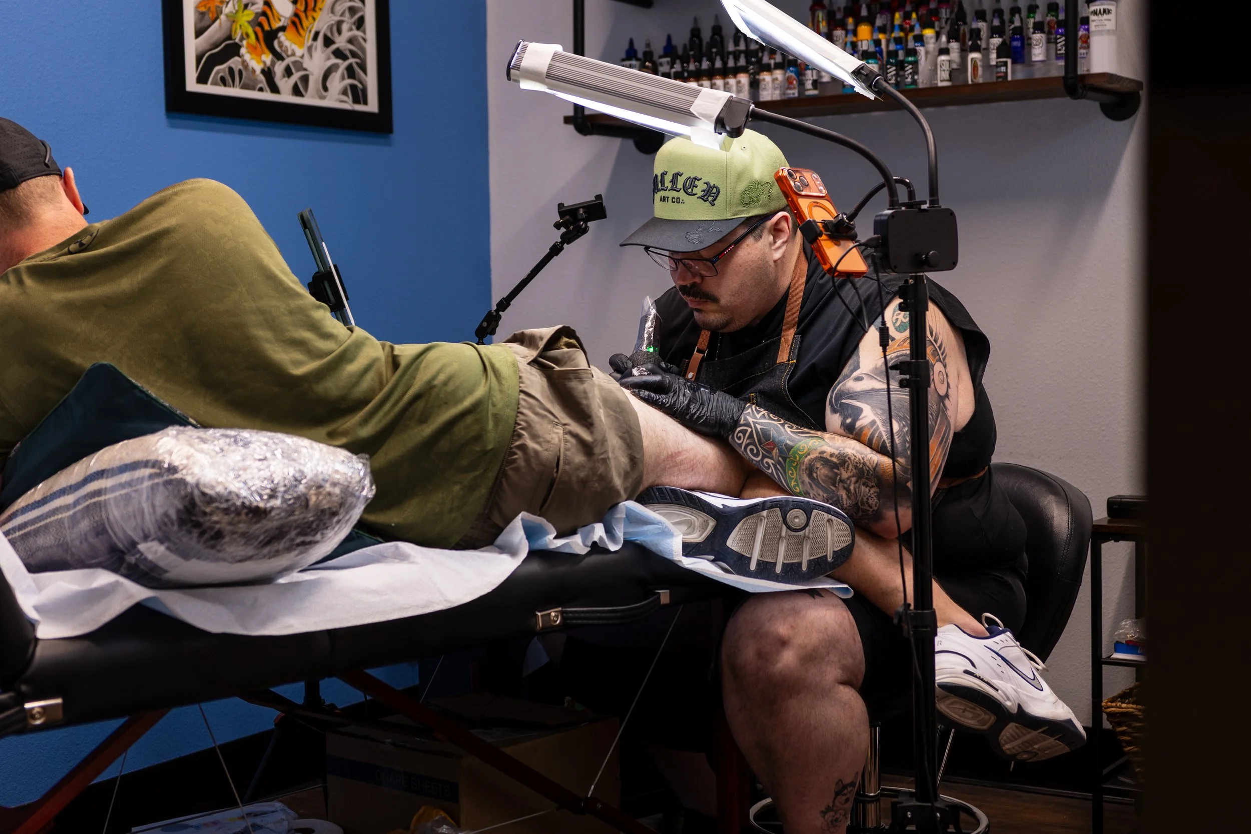 Tattoo artist tattooing a person's leg in a tattoo studio with blue walls, artwork, and ink bottles in the background.