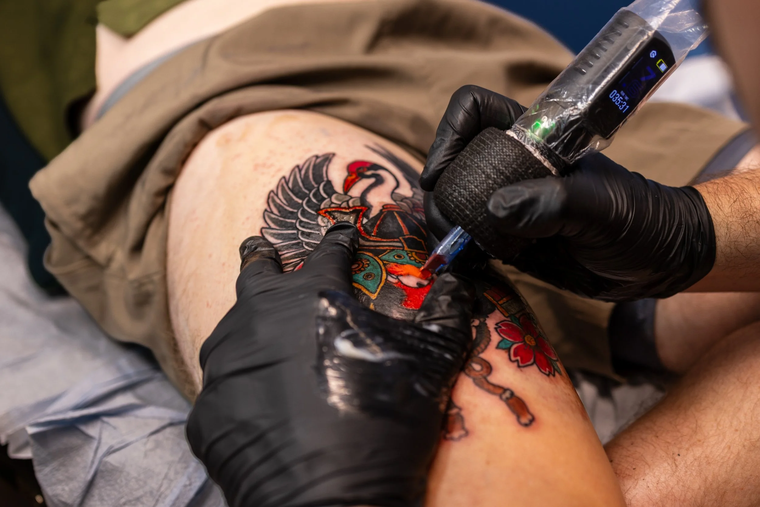 A person with tattoos on their thigh getting a tattoo done by a tattoo artist wearing black gloves.