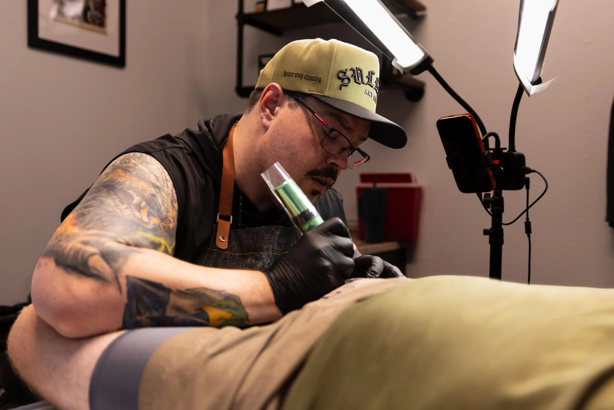 A man with tattoos on his arm wearing glasses, a beige cap with text, a black apron, and black gloves working on a tattoo or art project in a room with a lamp and a smartphone on a stand.