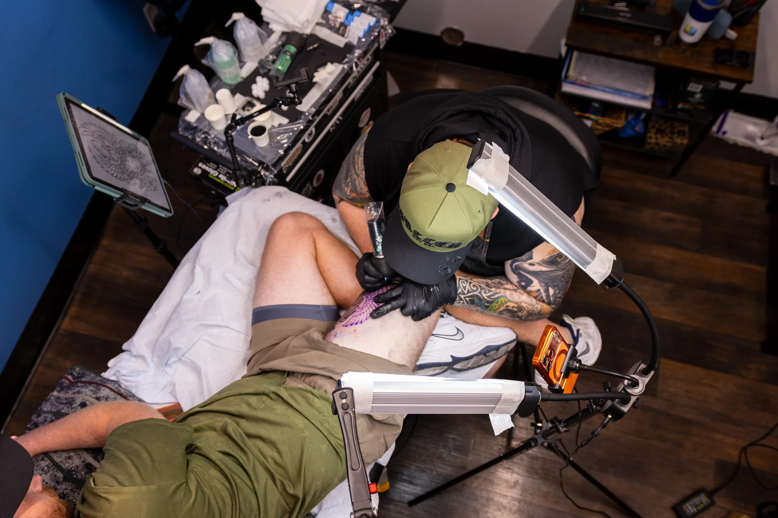 A person receiving a tattoo on their thigh from a tattoo artist in a tattoo studio. The artist is wearing a black shirt and green cap, and is using tattoo equipment. The client is laying down on a white sheet, wearing beige shorts and a green shirt.