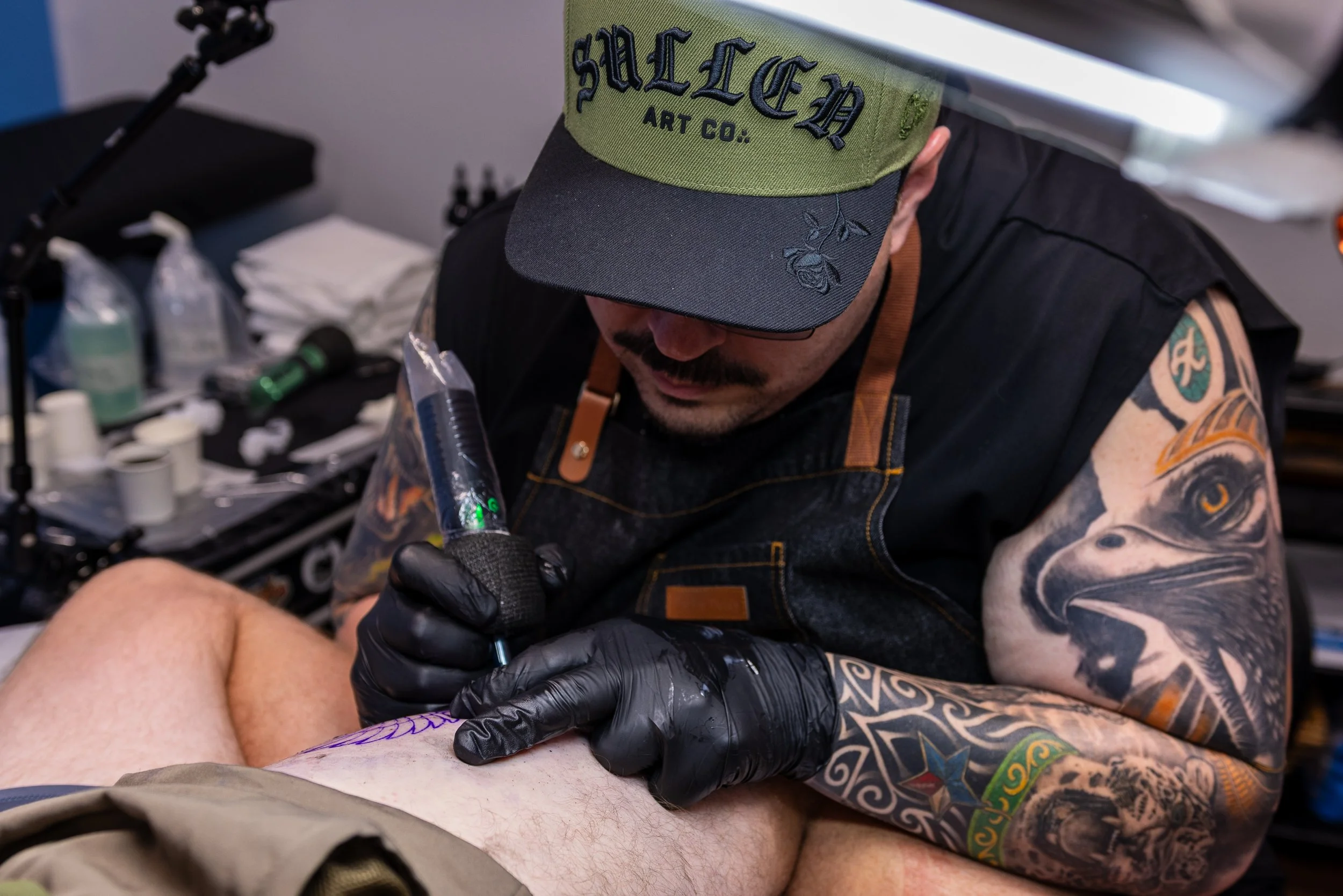 A tattoo artist with tattoos on his arms, wearing a black cap and gloves, tattooing a person's leg in a tattoo studio. The artist is focused on his work, surrounded by tattoo equipment and supplies.