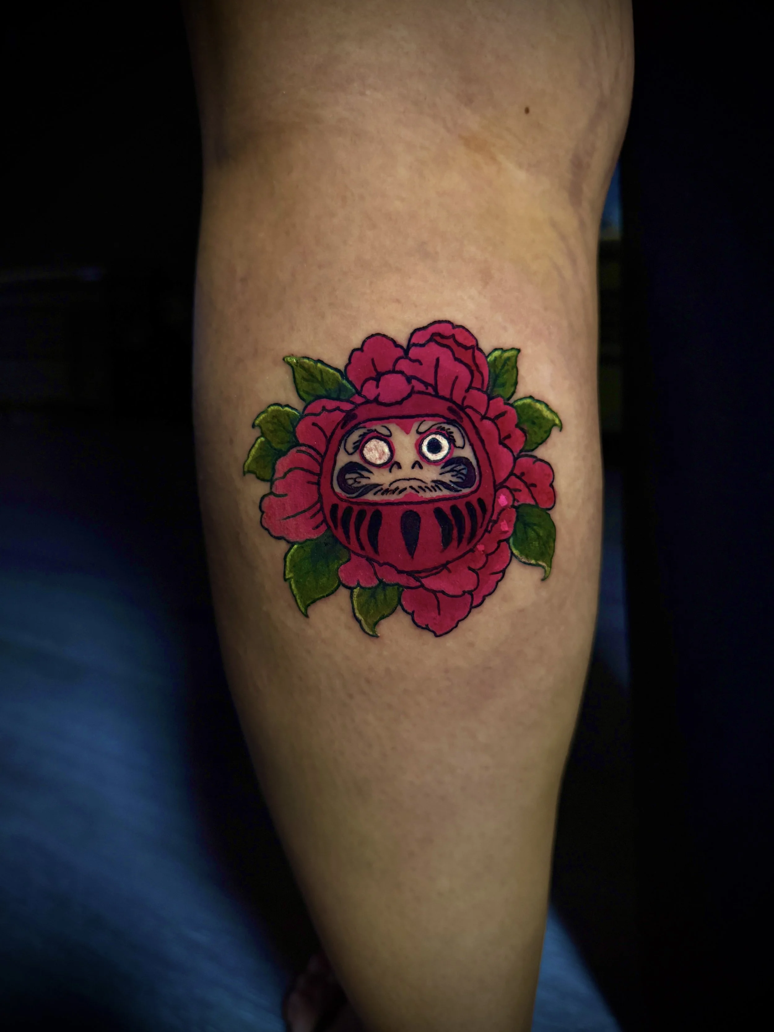 Tattoo of a Daruma doll with pink flowers and green leaves on a person's arm.