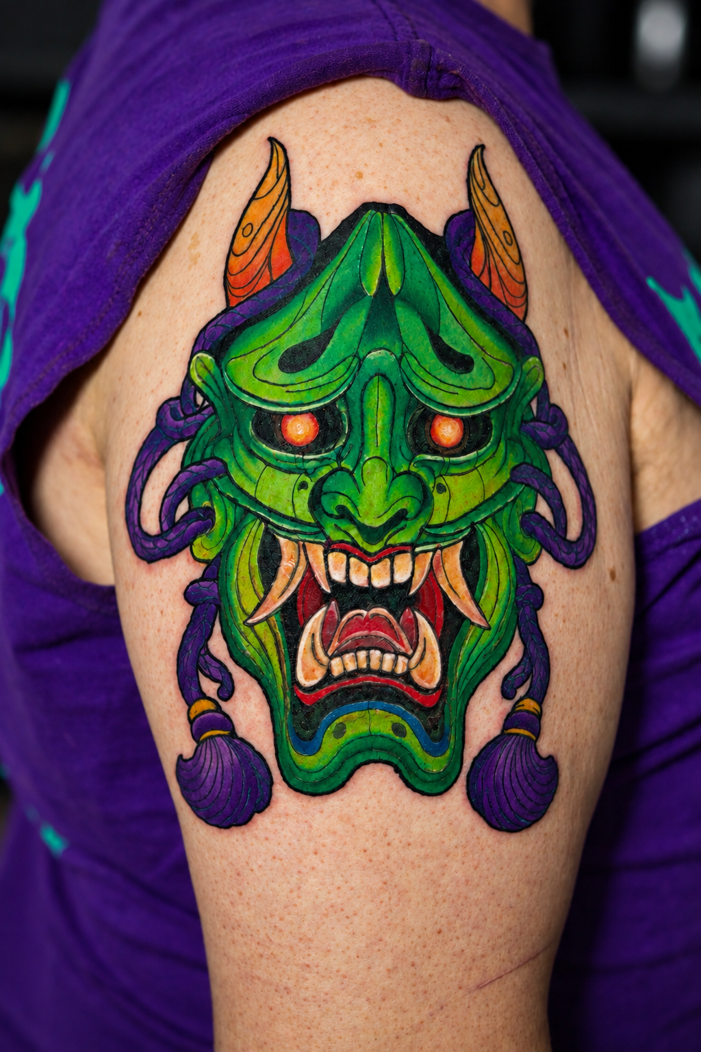 Colorful tattoo of a fierce green dragon or demon face with orange eyes, sharp teeth, and purple and orange accents on the shoulder of a person wearing a purple shirt.