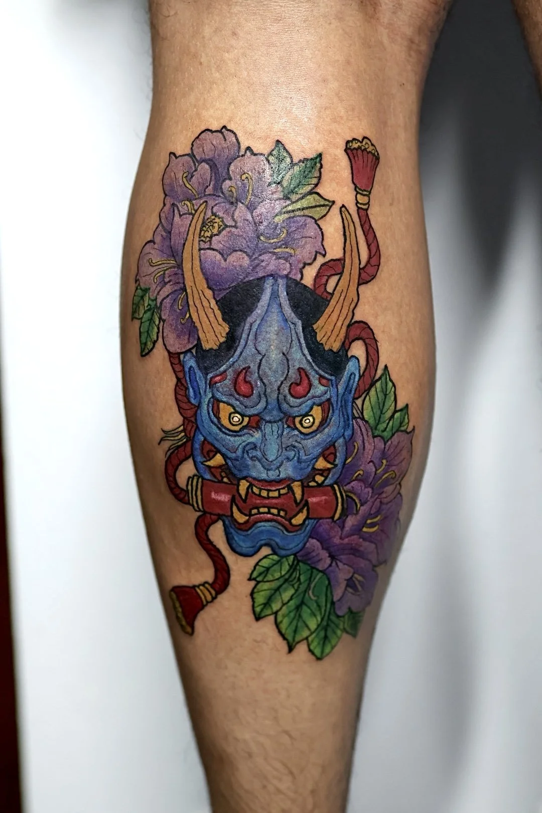 Colorful tattoo of a blue demon mask with red and yellow accents, horns, surrounded by purple flowers and green leaves.