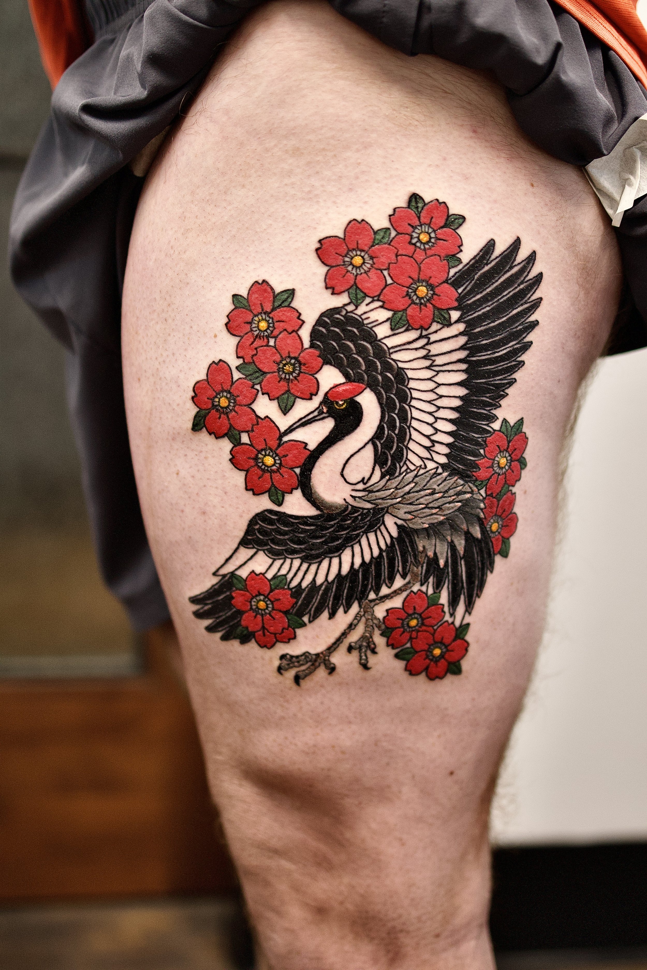 A tattoo of a black and white bird with outstretched wings and a long, slender beak, surrounded by red flowers with green leaves on a person's upper thigh.
