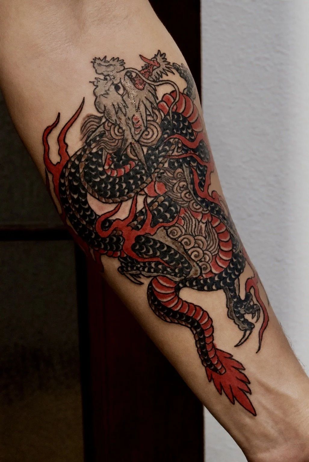 Tattoo of a fierce dragon with black and red scales, claws, and a long tail, coiled around the arm.