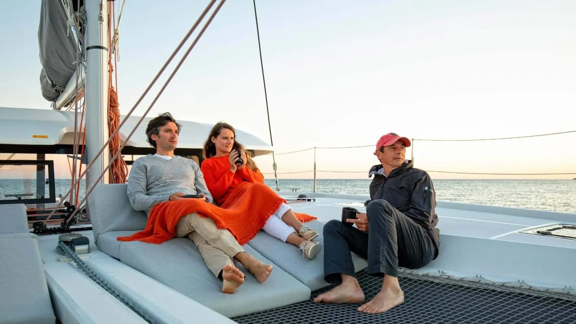 Three people sitting on a boat deck, looking at the water in the distance during sunset, with two of them holding mugs and one person holding a small device.