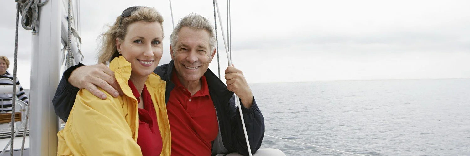 A smiling middle-aged man and woman on a boat, with the sea and cloudy sky in the background.