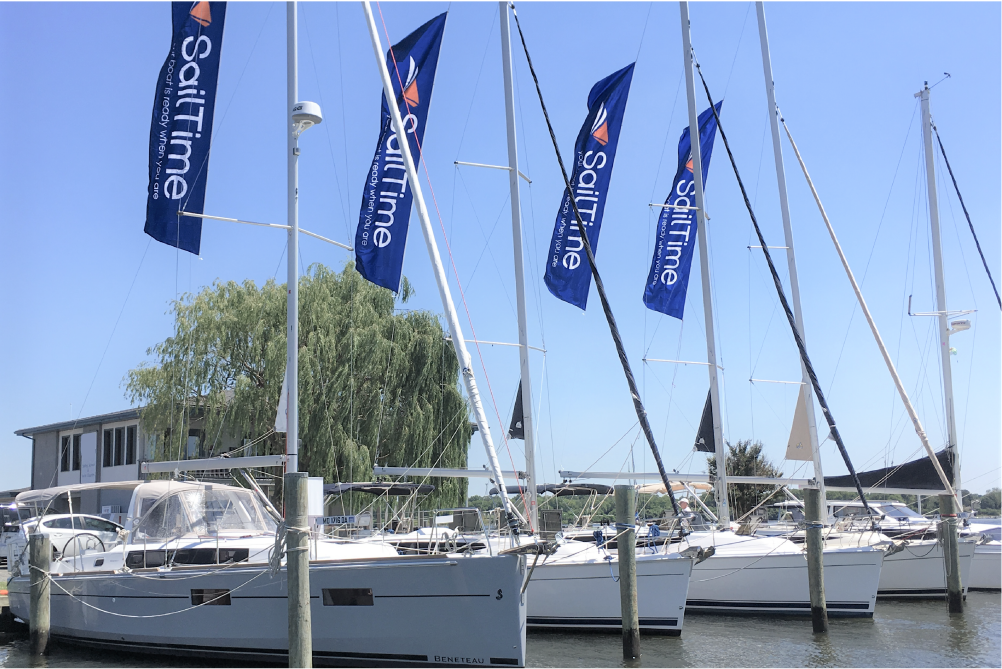 Several sailboats docked at a marina with blue flags that say 'SailTime' and a building with trees in the background under a clear sky.