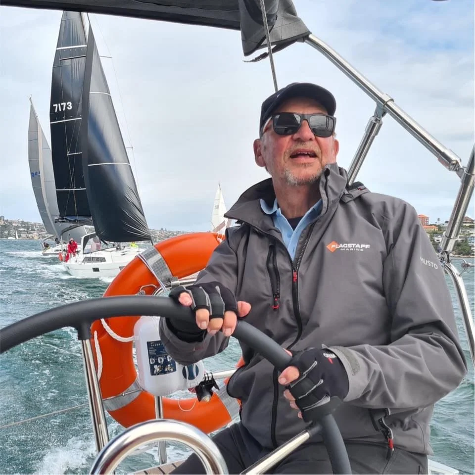 Man sailing a boat on the water, with other sailboats in the background, wearing sunglasses and a gray jacket.