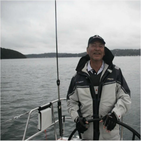 A man smiling on a boat in a body of water with distant land and overcast sky in the background.