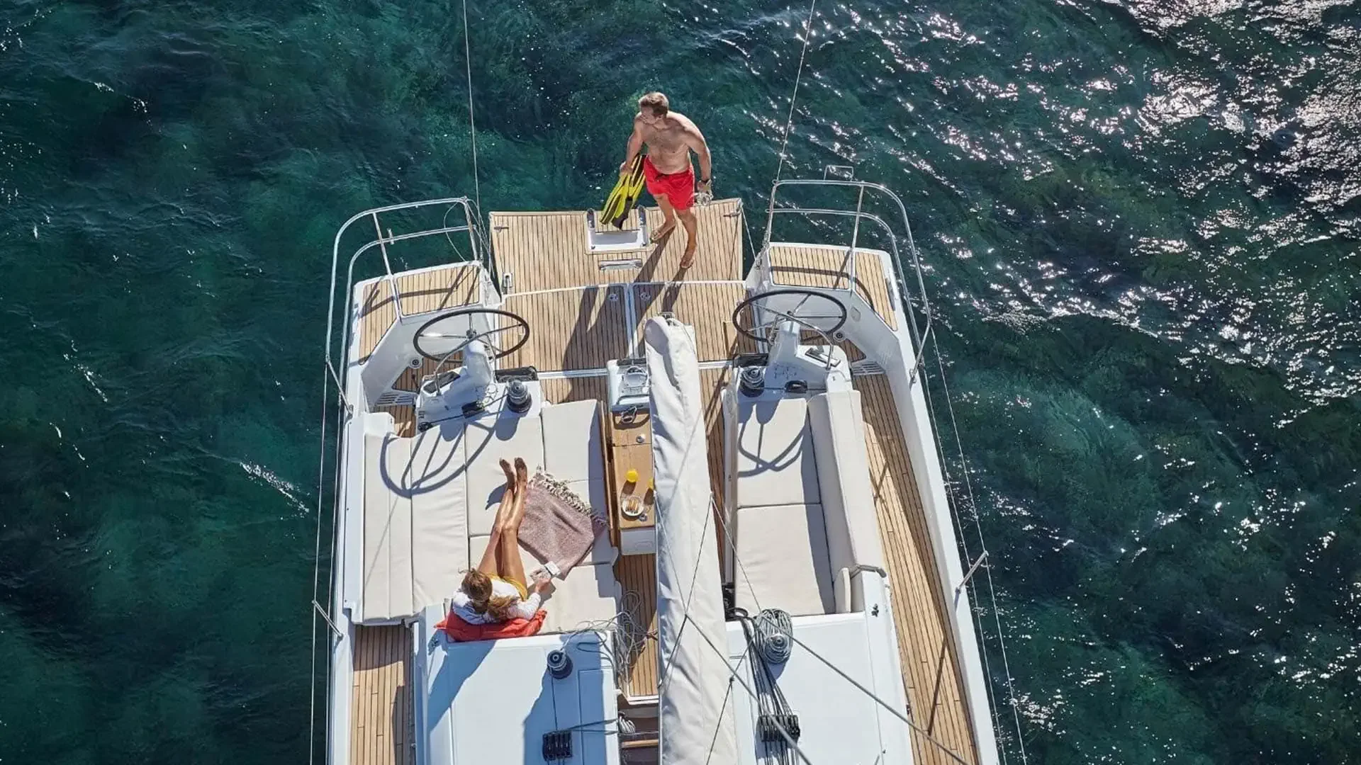 Aerial view of a sailboat on clear blue water with a man in red shorts and a woman with a towel and white top relaxing on the deck.