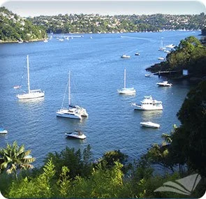 A river with sailboats and motorboats floating on it, bordered by green trees and hills in the background.