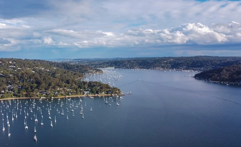 Aerial view of a large body of water with numerous sailboats docked along the shoreline and in the water, surrounded by green hilly terrain and a cloudy sky.