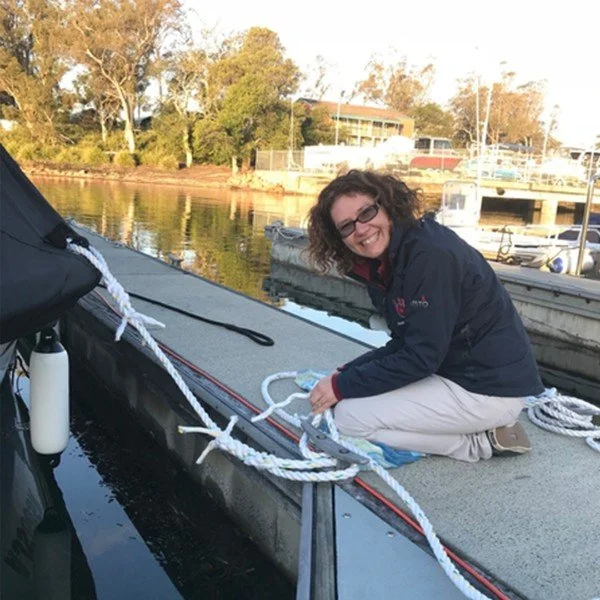 Woman with curly hair and glasses crouching on a dock near a sailboat, smiling at the camera, with a marina and trees in the background.