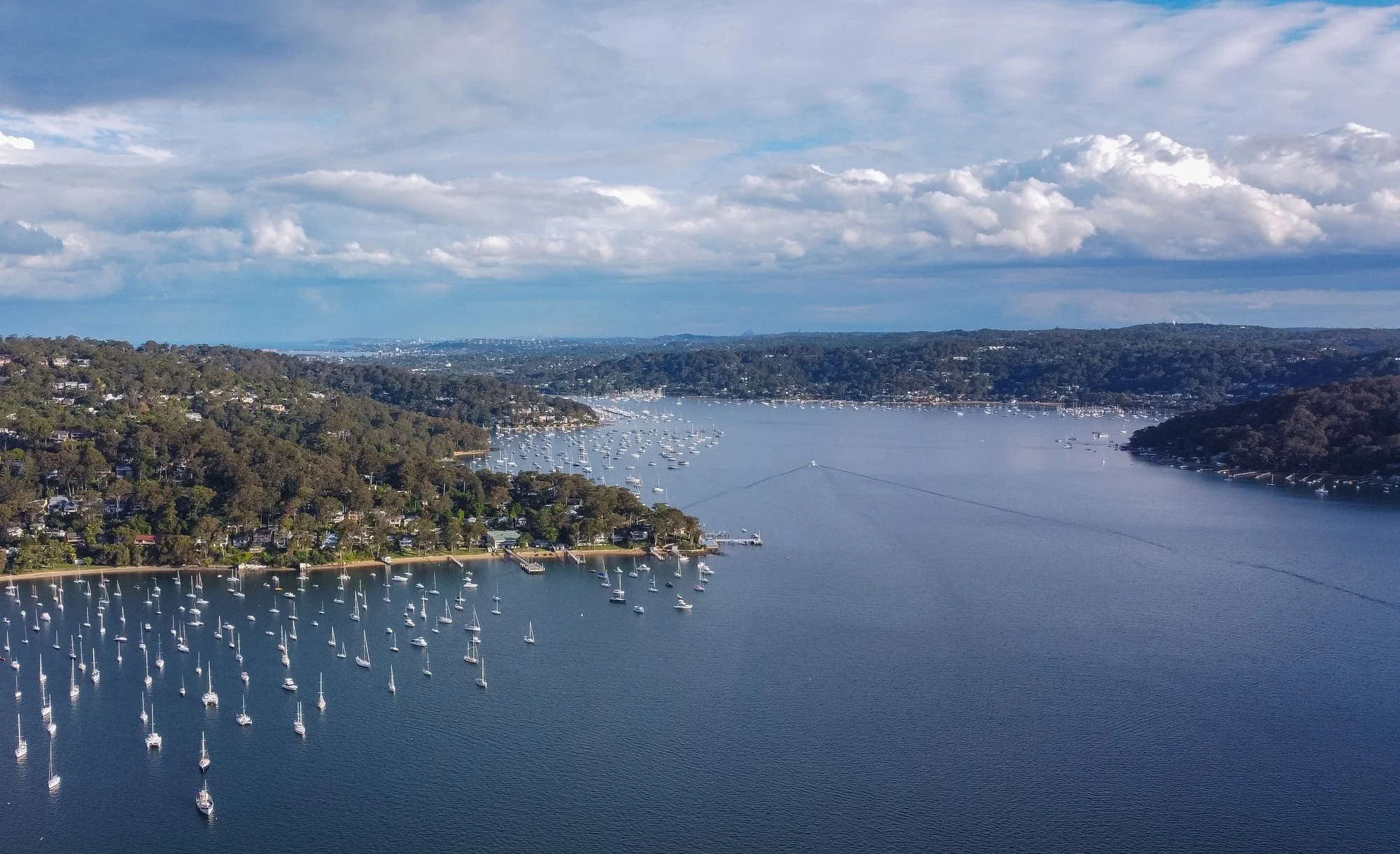 Aerial view of a large body of water with numerous sailboats anchored, surrounded by green hills and a cloudy sky.