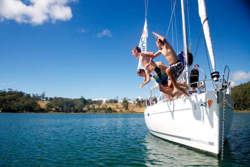 Three children jumping into the water from a white sailboat on a sunny day.