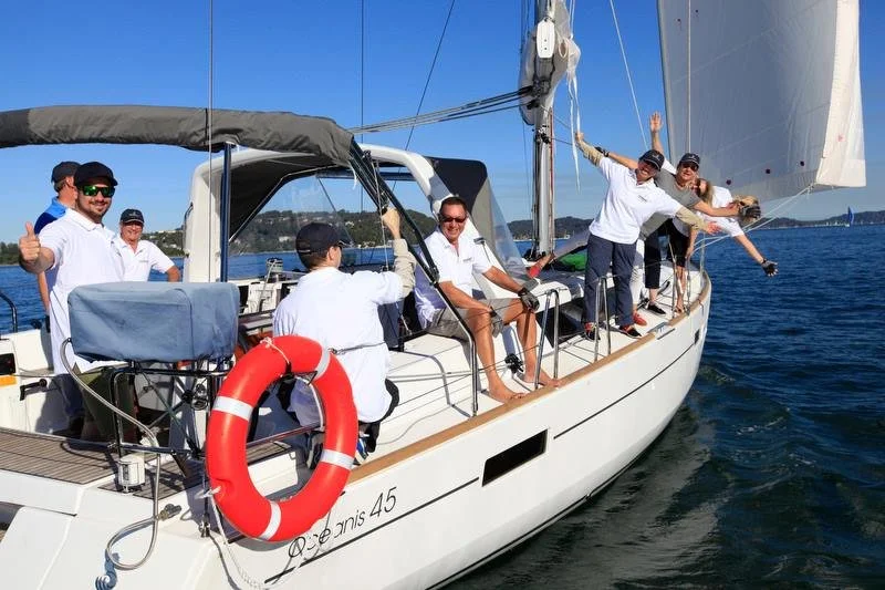 Group of people on a sailboat enjoying a day on the water, some waving and smiling, with a clear blue sky and calm sea in the background.