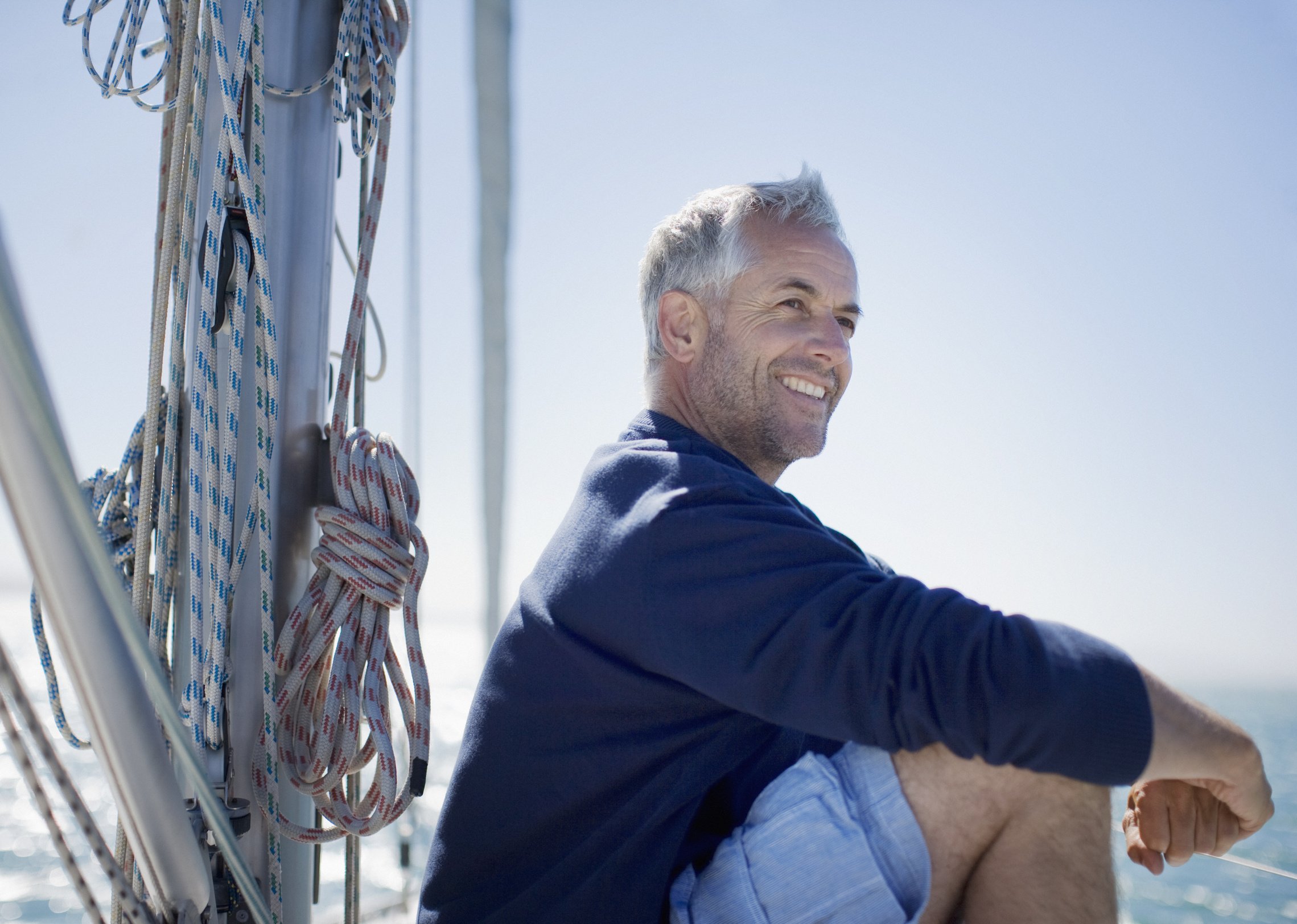 A man sitting on a boat with sailing ropes and equipment nearby, smiling and looking into the distance under a clear blue sky.