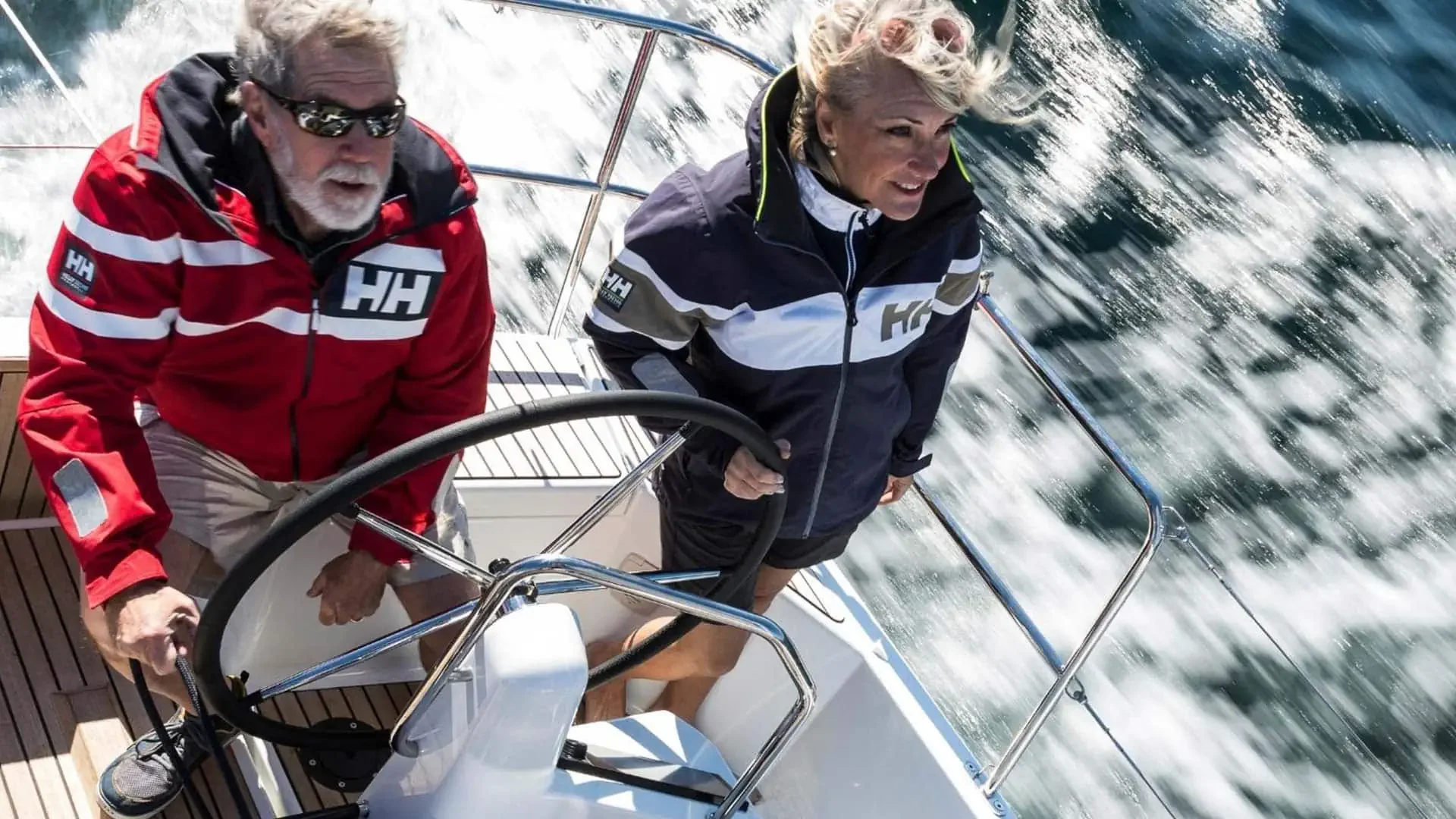Two people steering a sailing boat on the water, wearing Helly Hansen jackets, with waves and wake in the background.