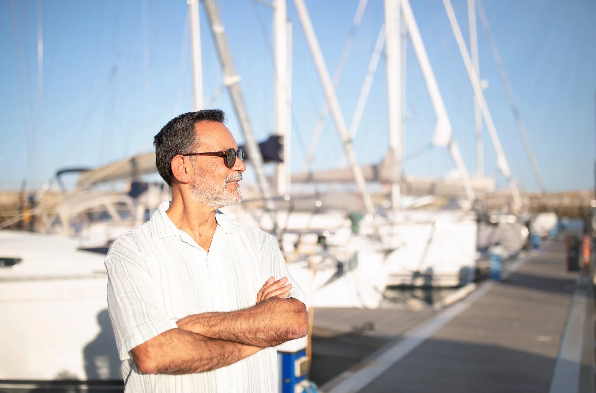 A middle-aged man with dark hair, gray beard, wearing sunglasses and a light-colored striped shirt, standing with arms crossed at a marina filled with sailboats under a clear blue sky.