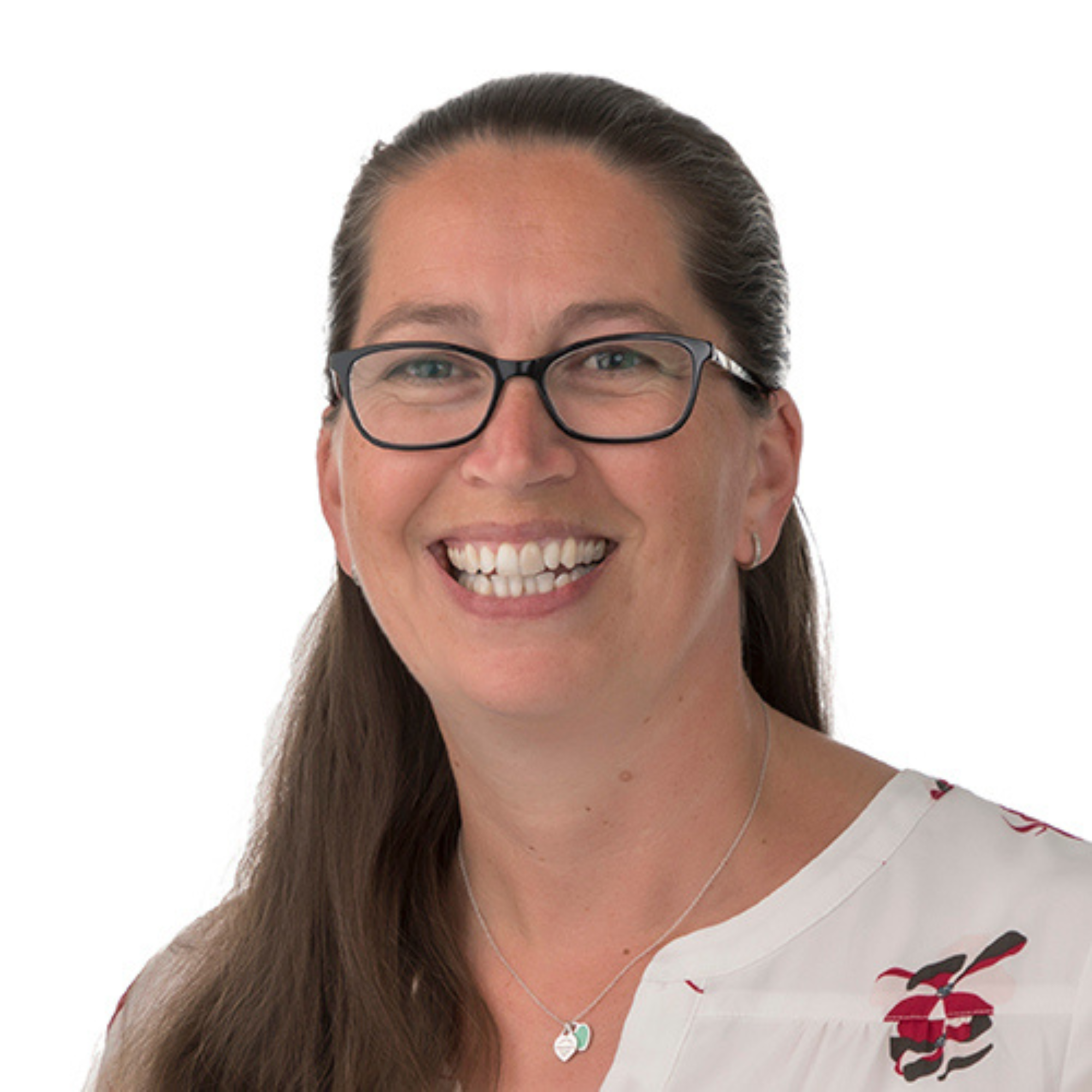 A woman with long brown hair, glasses, and a bright smile, wearing a white top with floral embroidery and a heart-shaped pendant necklace.