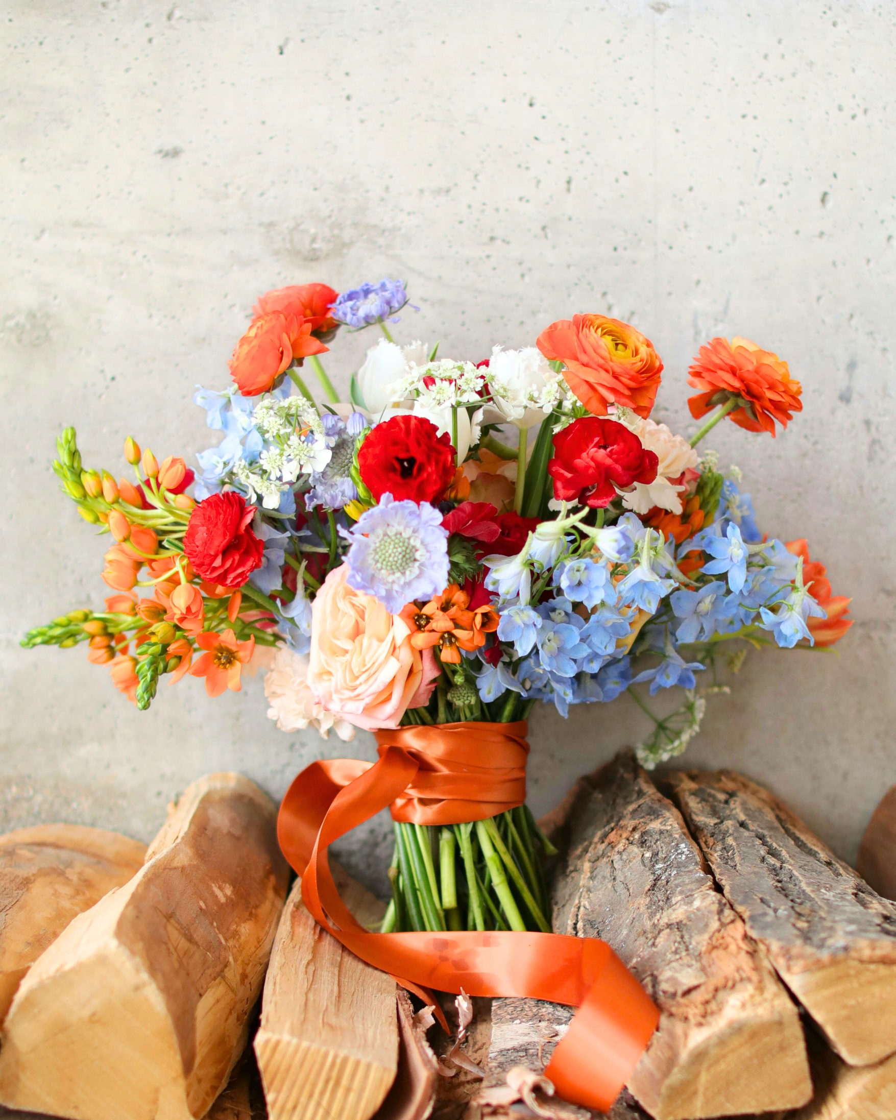 Colorful flower bouquet with orange, red, blue, and peach blooms, tied with an orange ribbon, placed on stacked firewood against a concrete wall.