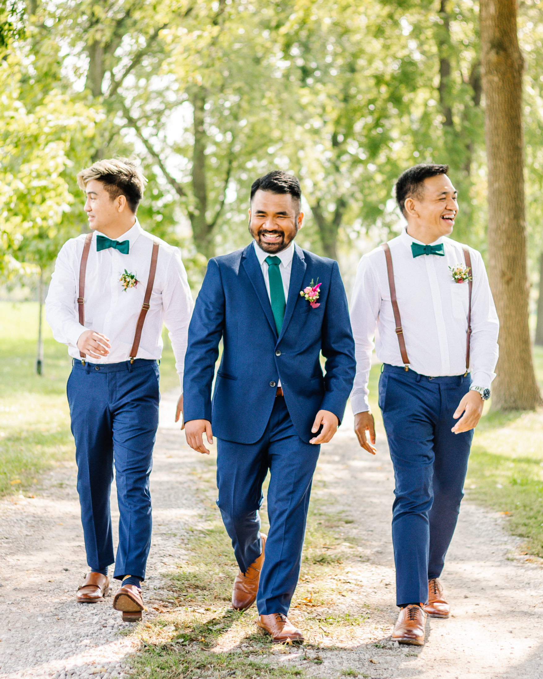 Three men walking outdoors in formal attire, wearing blue suits, white shirts, and bow ties. One man wears a full suit, while the other two wear suspenders. They are smiling and walking on a path surrounded by trees.