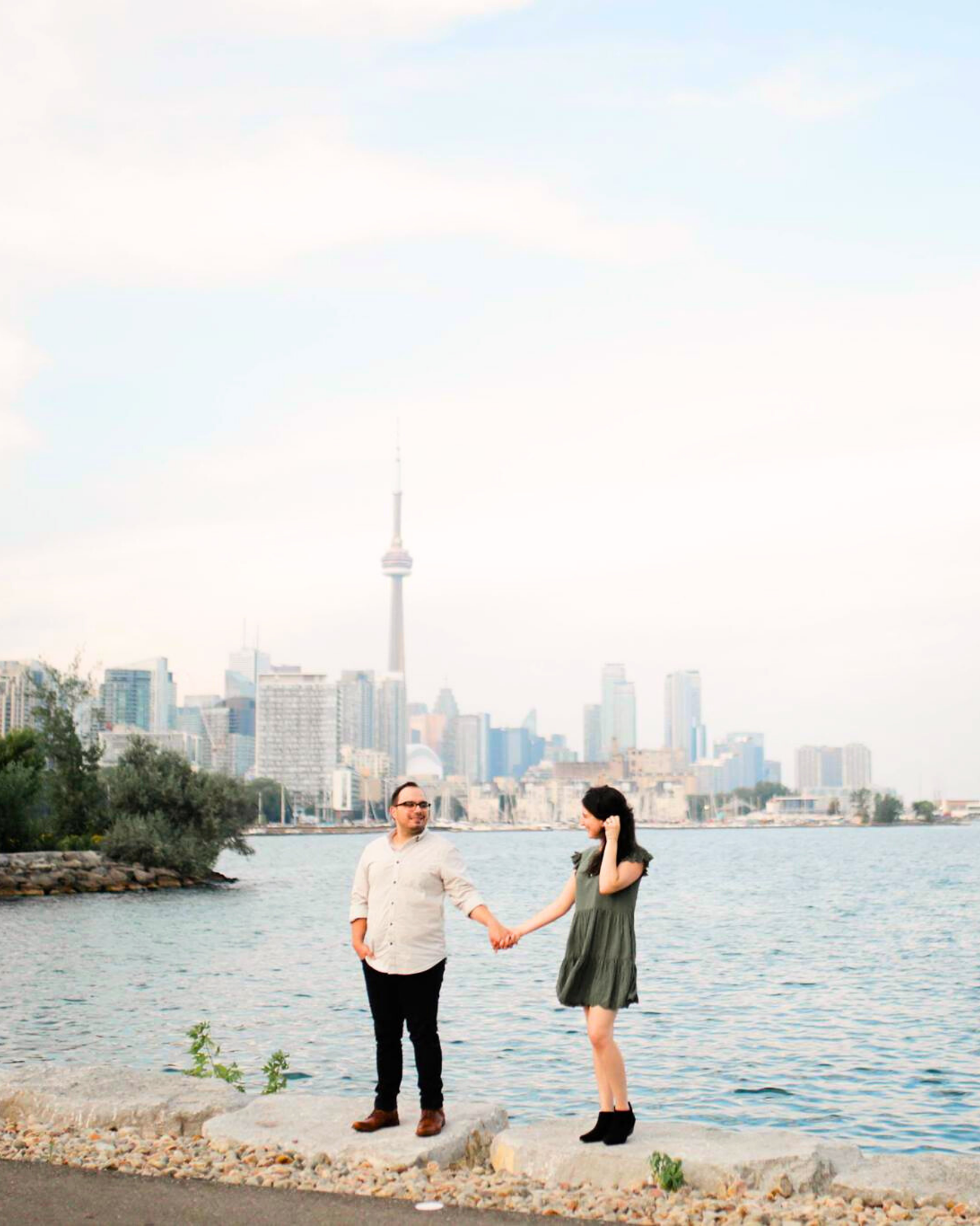 A couple holding hands on a waterfront with Toronto's skyline, including the CN Tower, in the background.