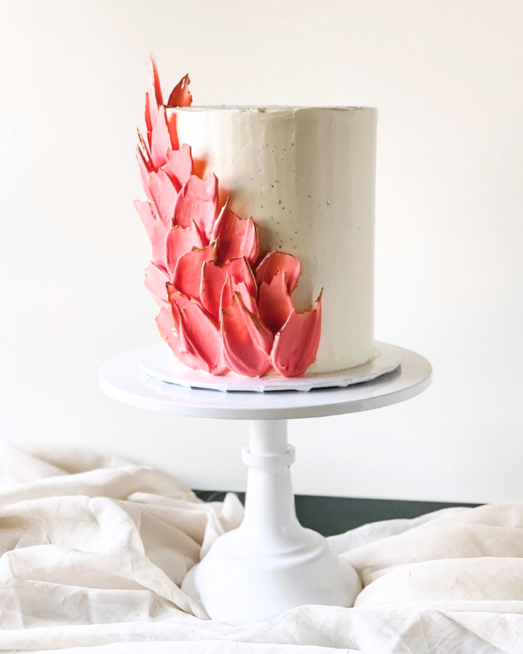 Tall white cake on a stand with pink petal decorations.