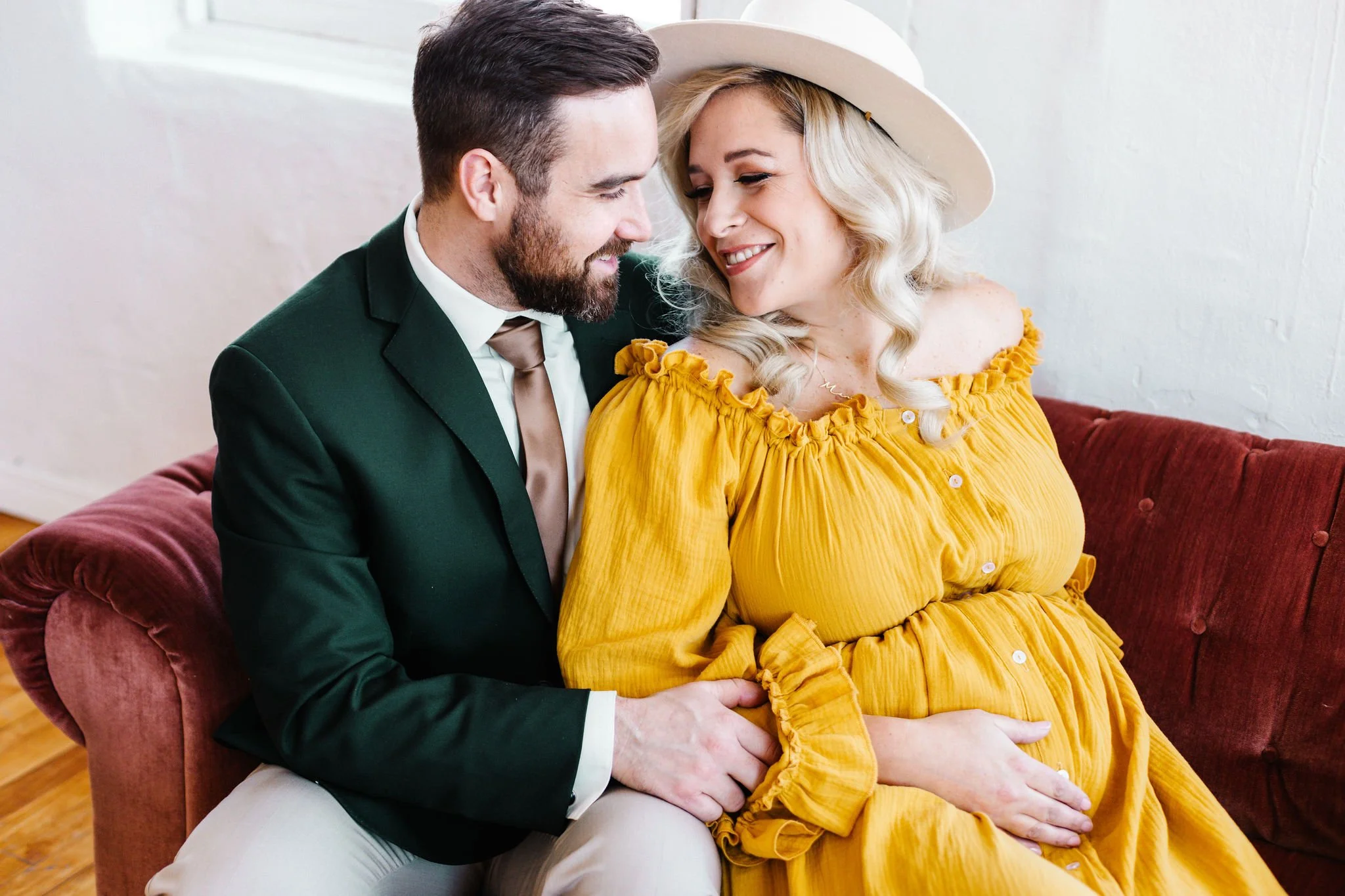 A smiling couple sitting on a sofa, the woman wearing a yellow dress and a hat, with the man's hand on her knee.