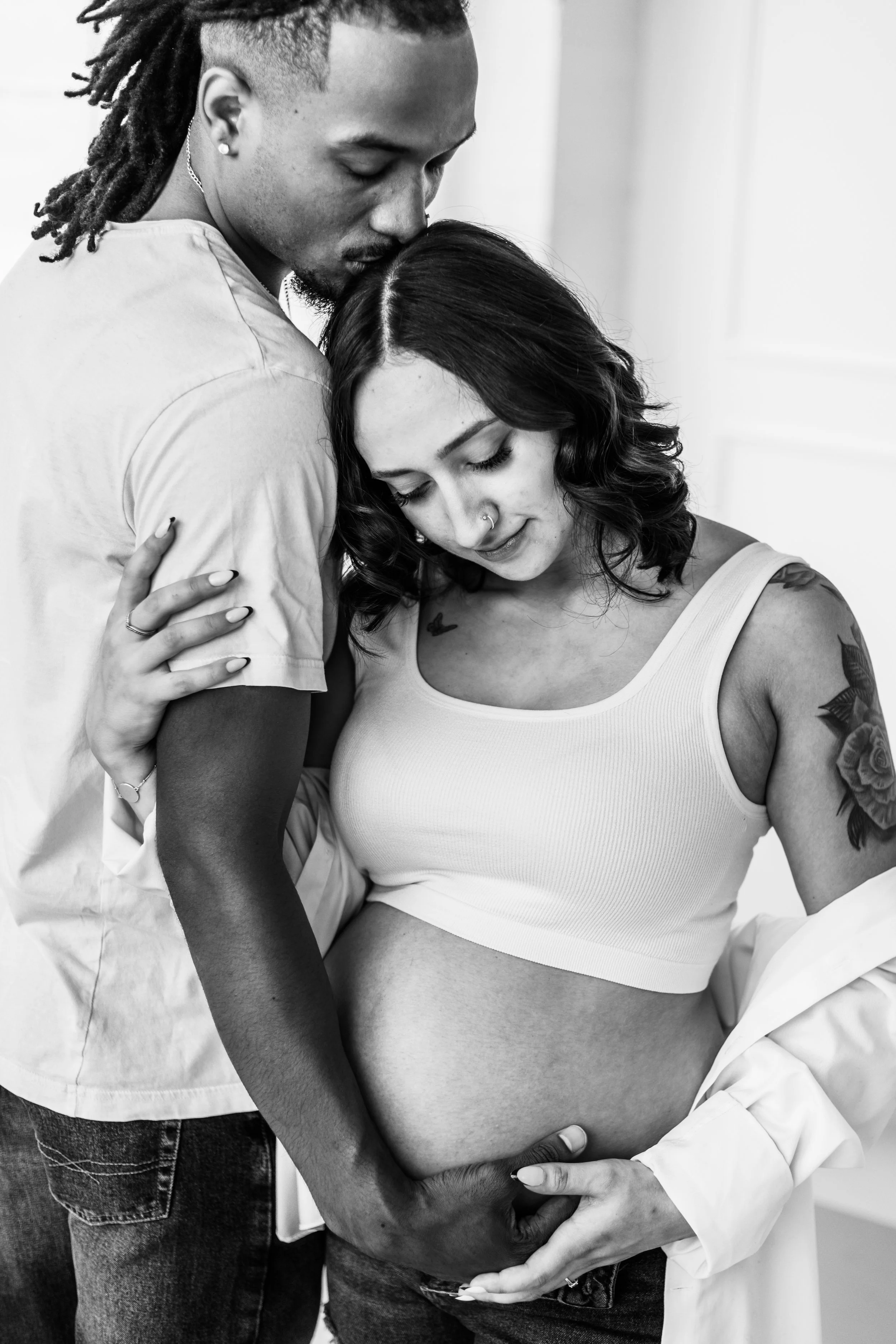 Black and white photo of an affectionate couple embracing, with the woman showing a bare pregnant belly. The man is holding the woman's belly and kissing her head.