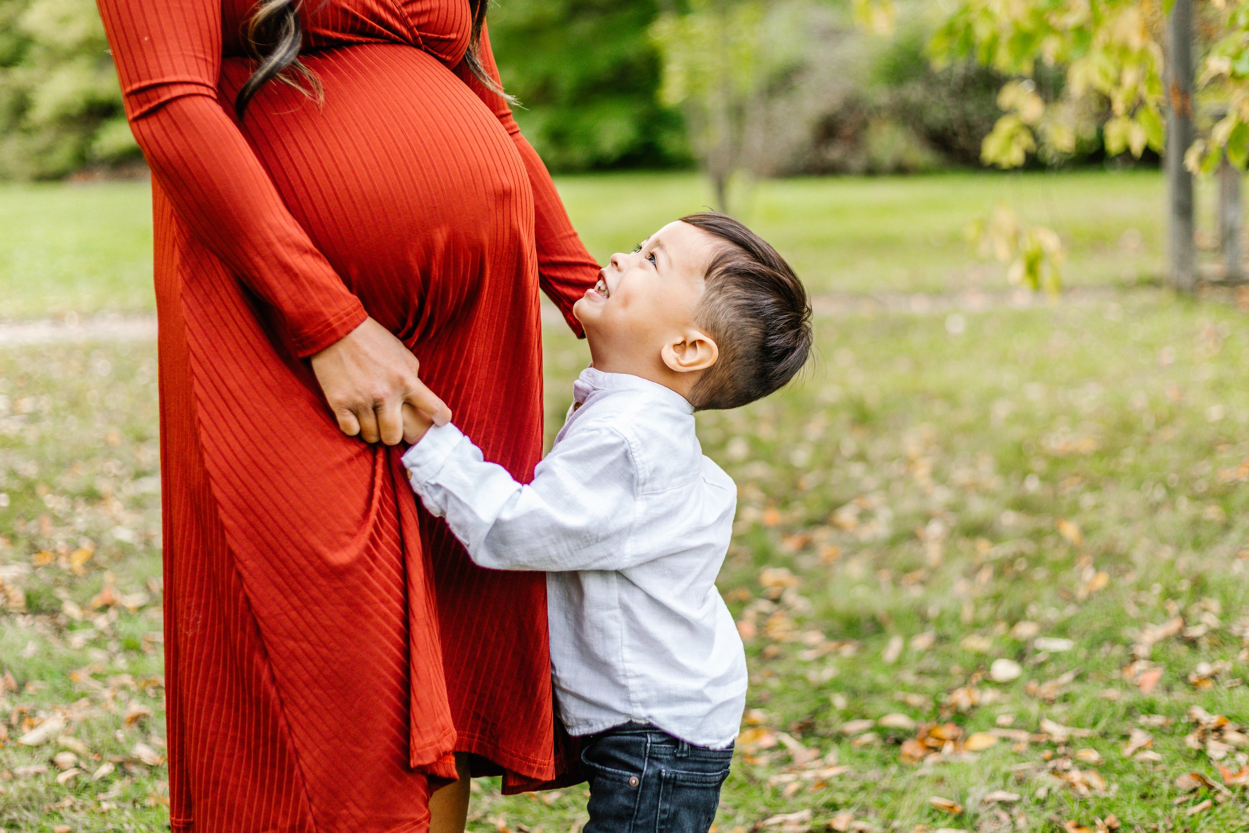 A pregnant woman in a red dress stands holding hands with a young boy in a white shirt. The child looks up, smiling at the woman, in an outdoor setting with grass and trees in the background.