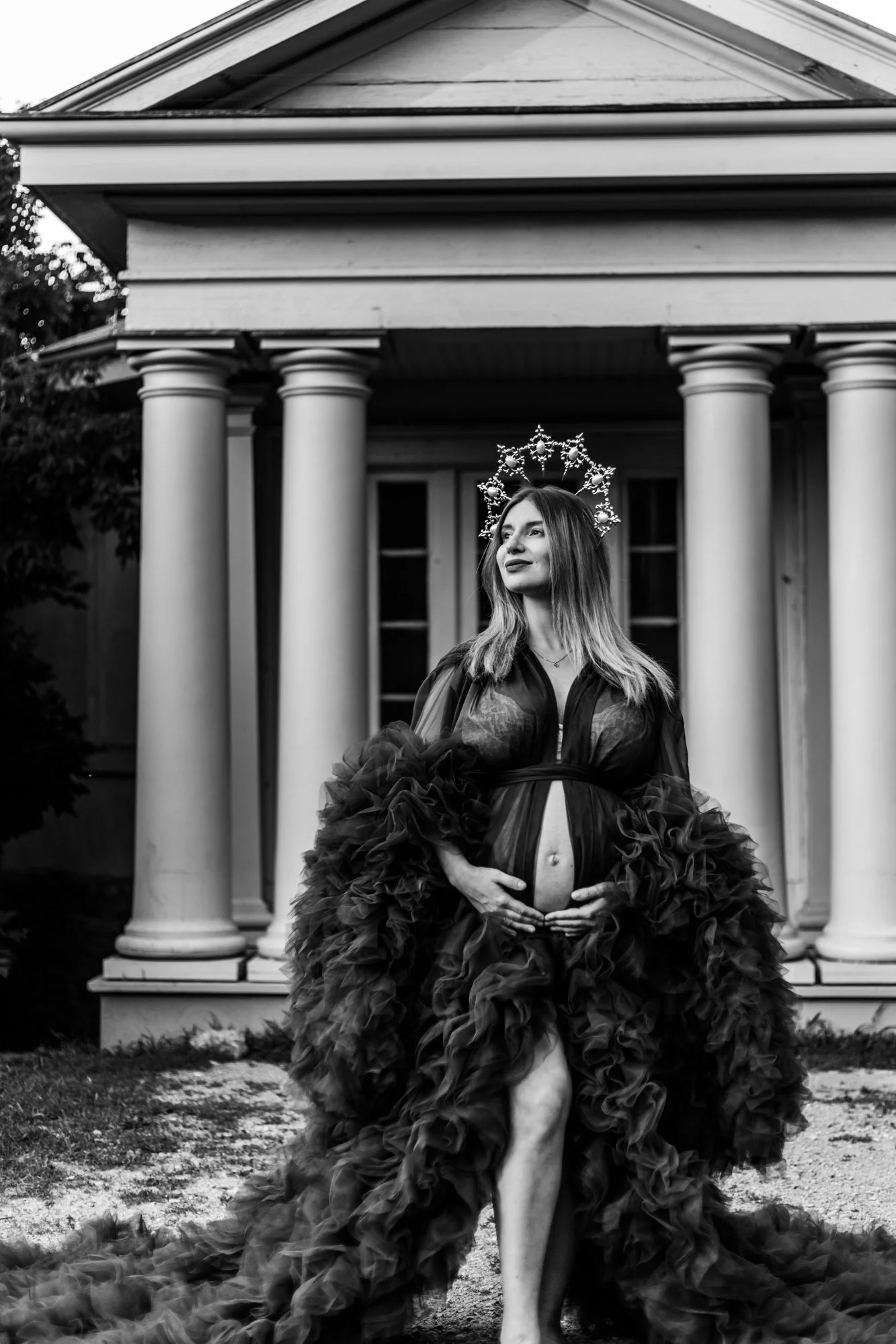 Pregnant woman in an elegant gown with a crown, standing in front of columns.