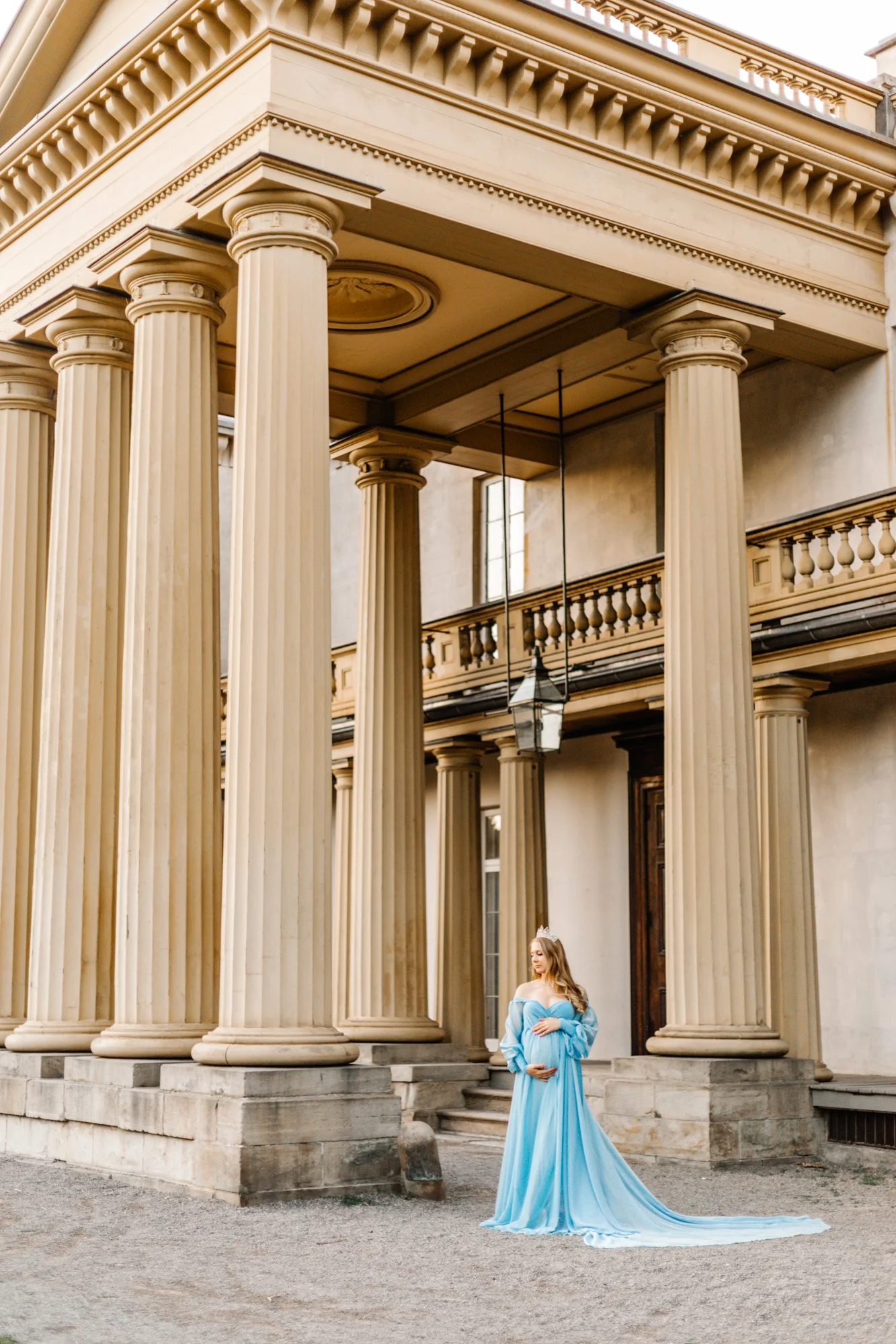 A woman in a flowing blue gown standing in front of a neoclassical building with large columns, holding her pregnant belly.
