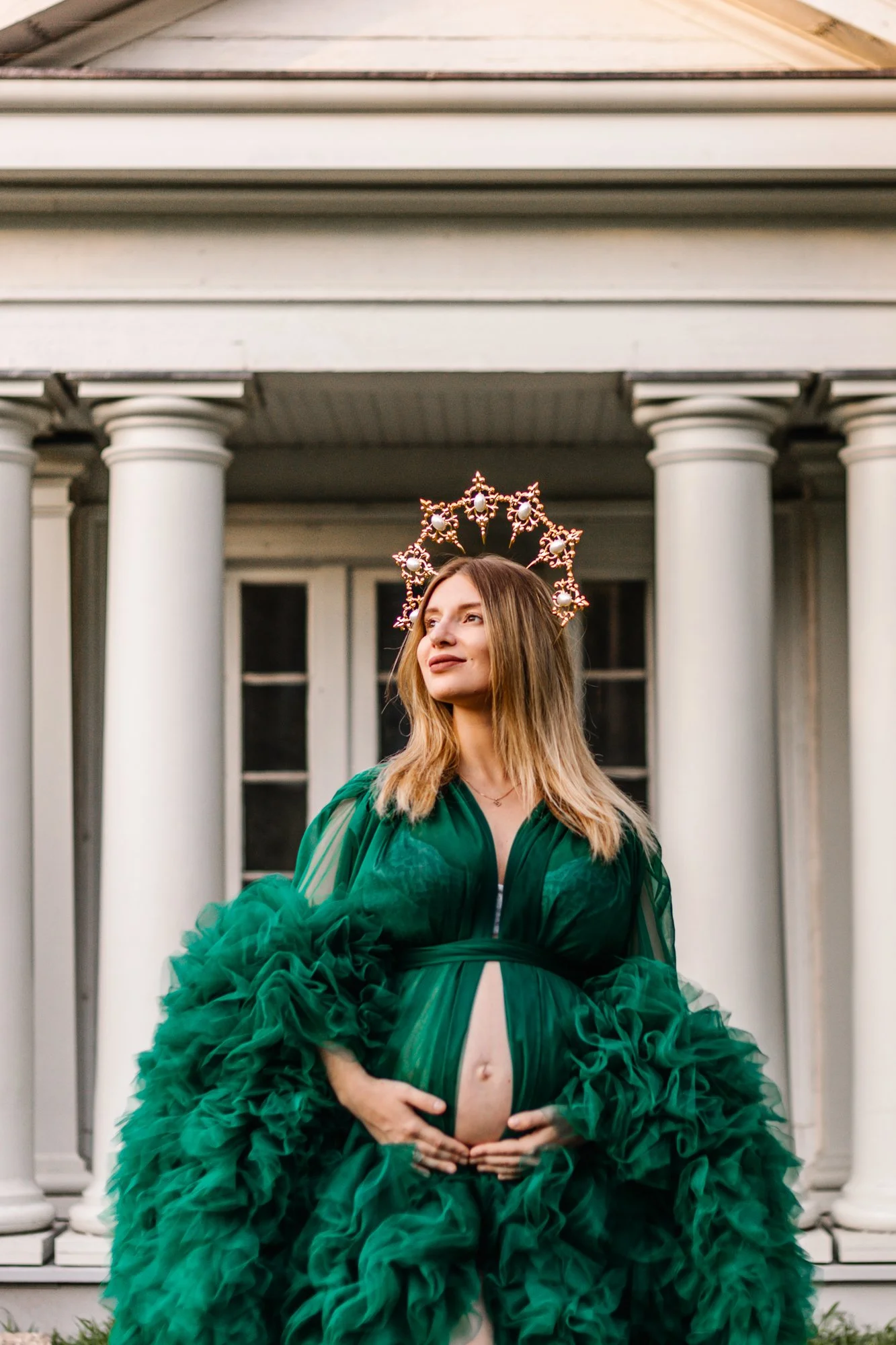 Pregnant woman in a green gown with a floral crown standing in front of white pillars.