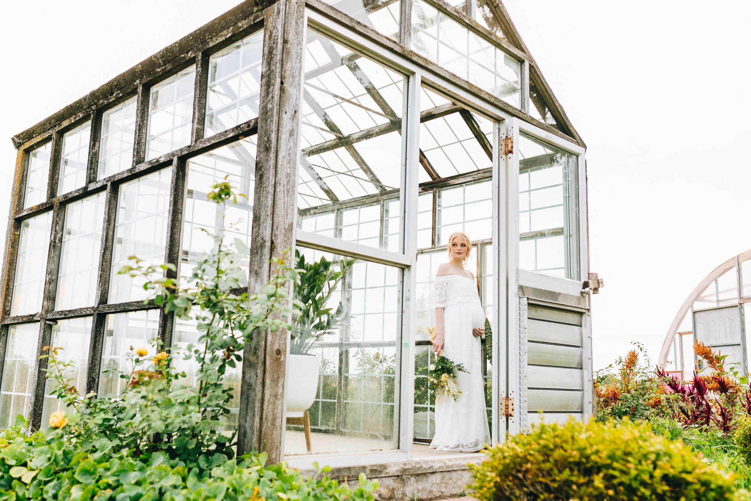 Bride in white dress standing in rustic wooden greenhouse surrounded by plants and flowers.