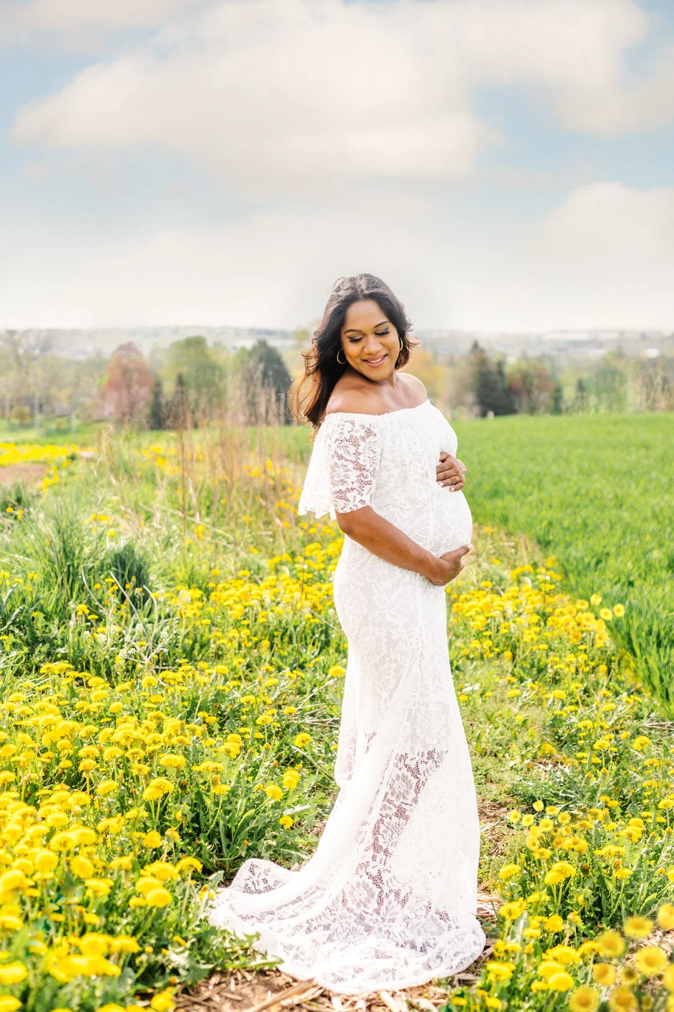 Pregnant woman in white lace dress standing in a field of yellow flowers, with a serene landscape in the background.