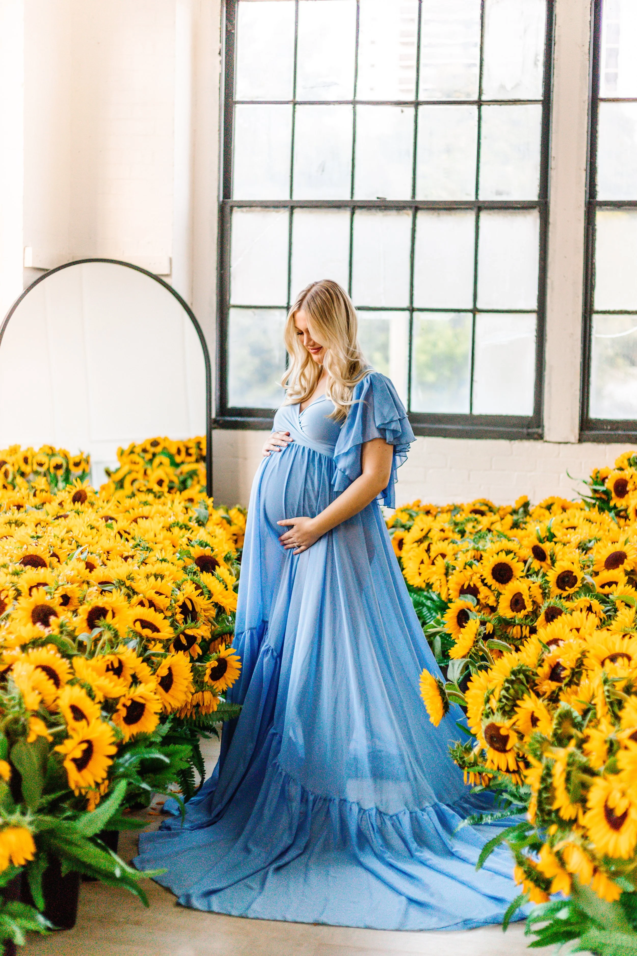 Pregnant woman in blue dress standing among numerous sunflowers, with a mirror reflecting the scene, inside a room with large grid windows.