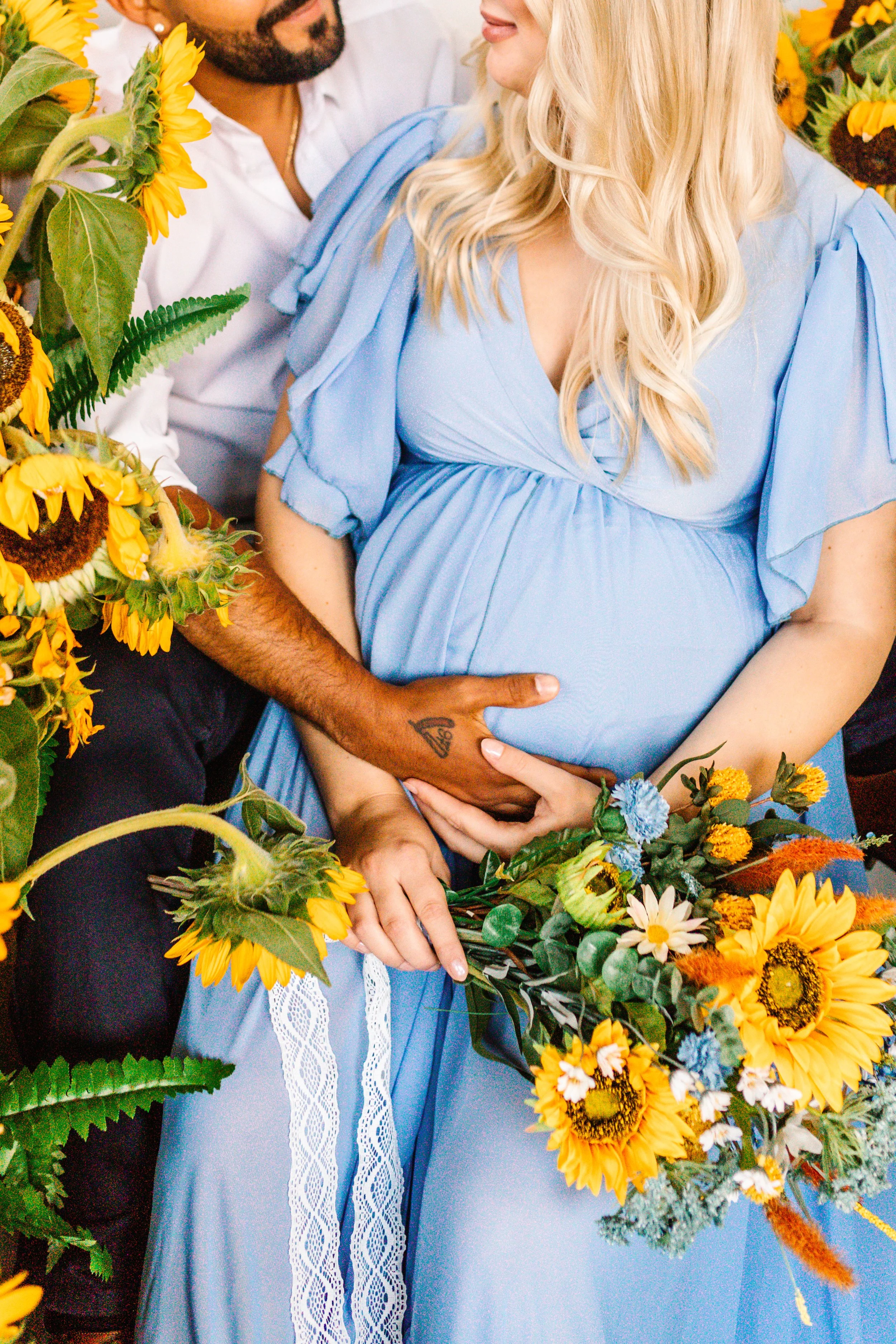 A pregnant woman in a blue dress is sitting amidst sunflowers, holding a bouquet. Her partner, wearing a white shirt, embraces her, placing a hand on her belly.