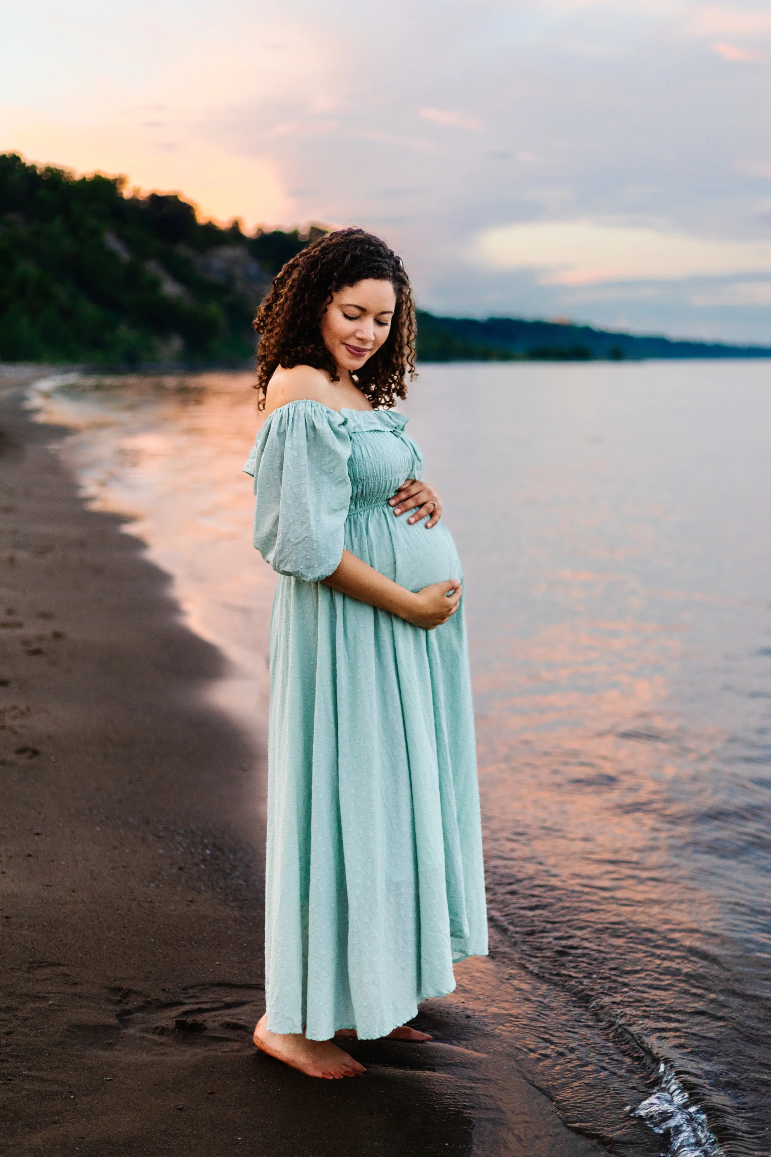 Pregnant woman in a light turquoise dress standing on a beach at sunset.