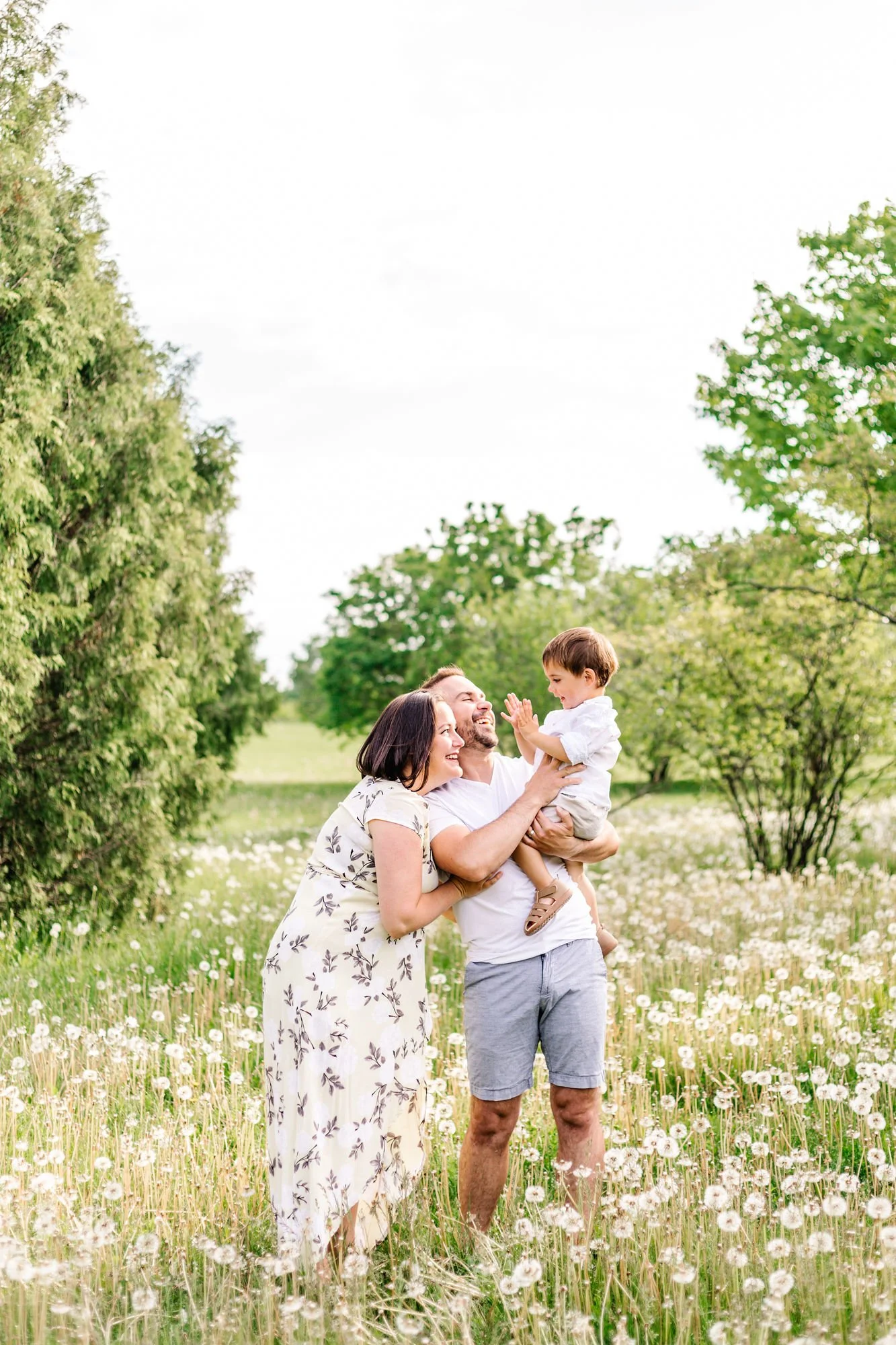 A happy family of three enjoying time in a field of dandelions with trees in the background. The mother and father are smiling and standing together while holding their child, who is joyfully playing and reaching out.