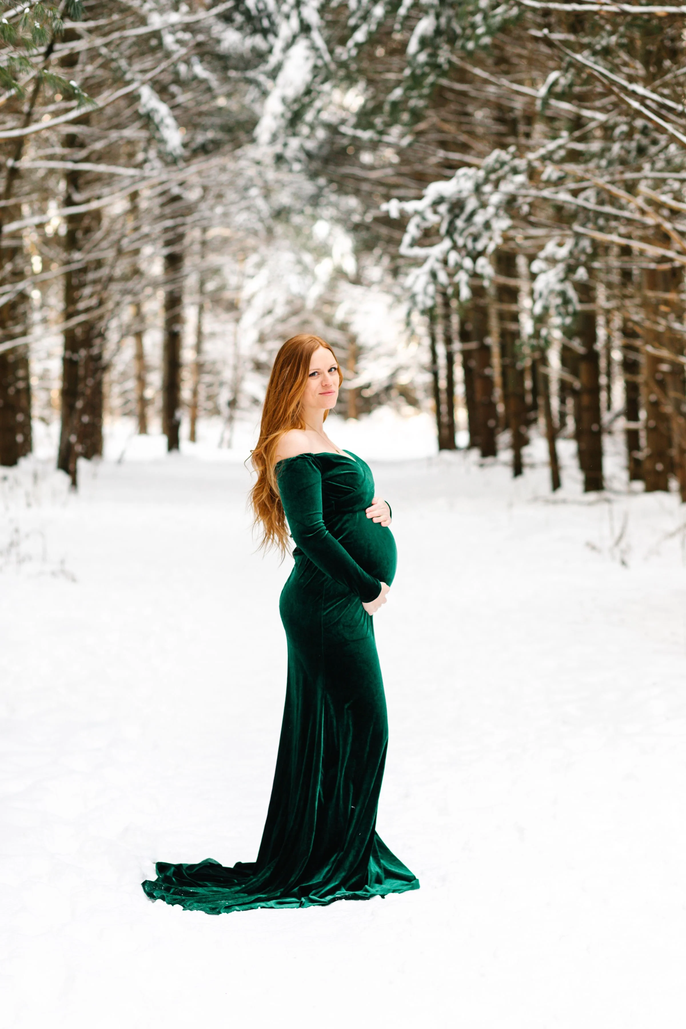 Pregnant woman in green dress standing in snowy forest.