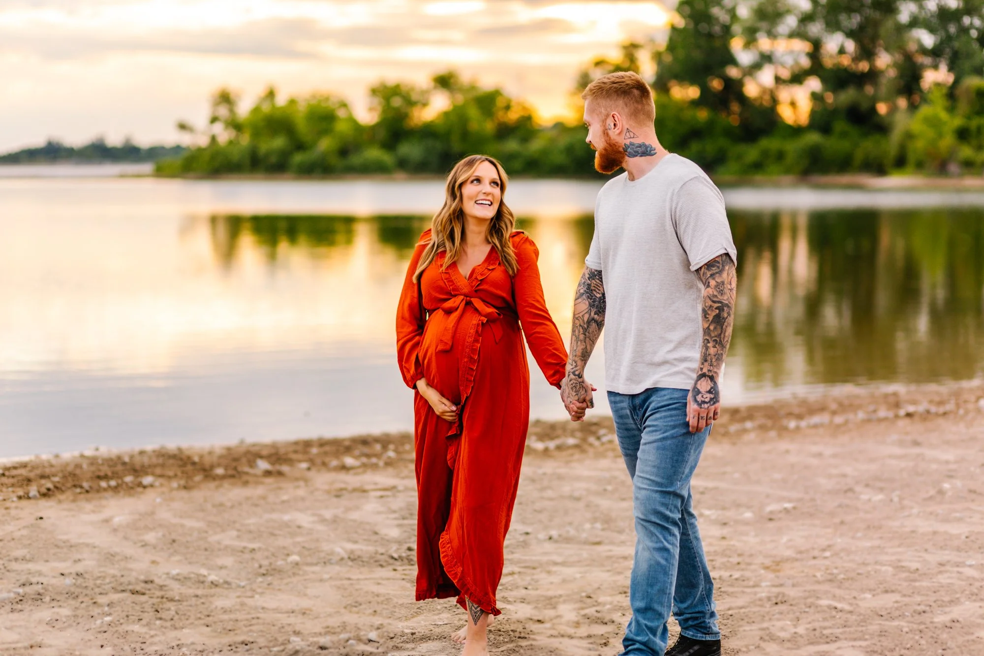 Pregnant woman in a red dress holding hands with a tattooed man by a lake at sunset.
