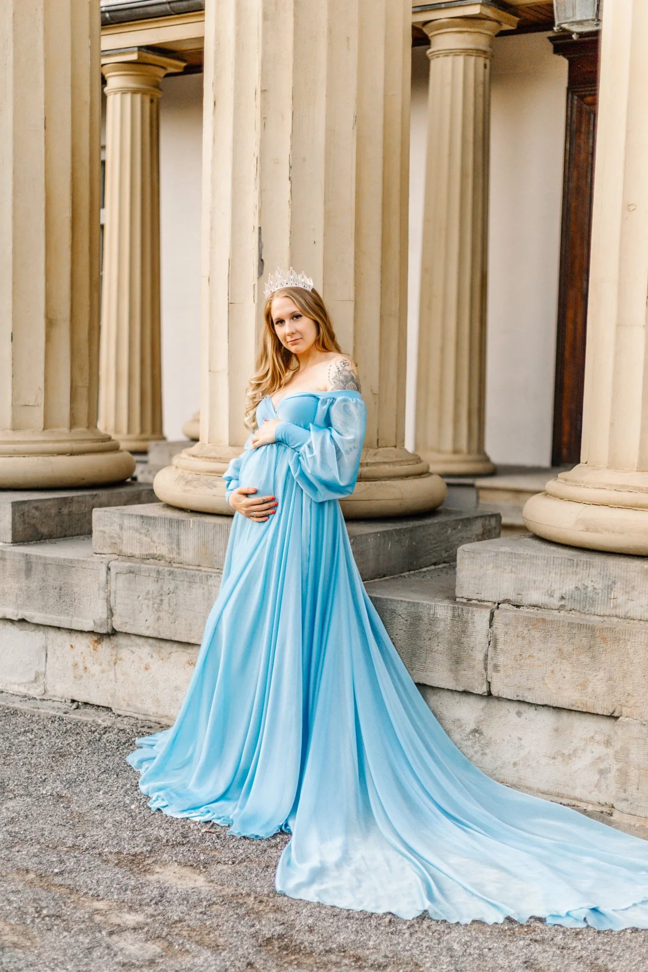 A pregnant woman wearing a flowing blue gown and a tiara poses in front of stone columns.