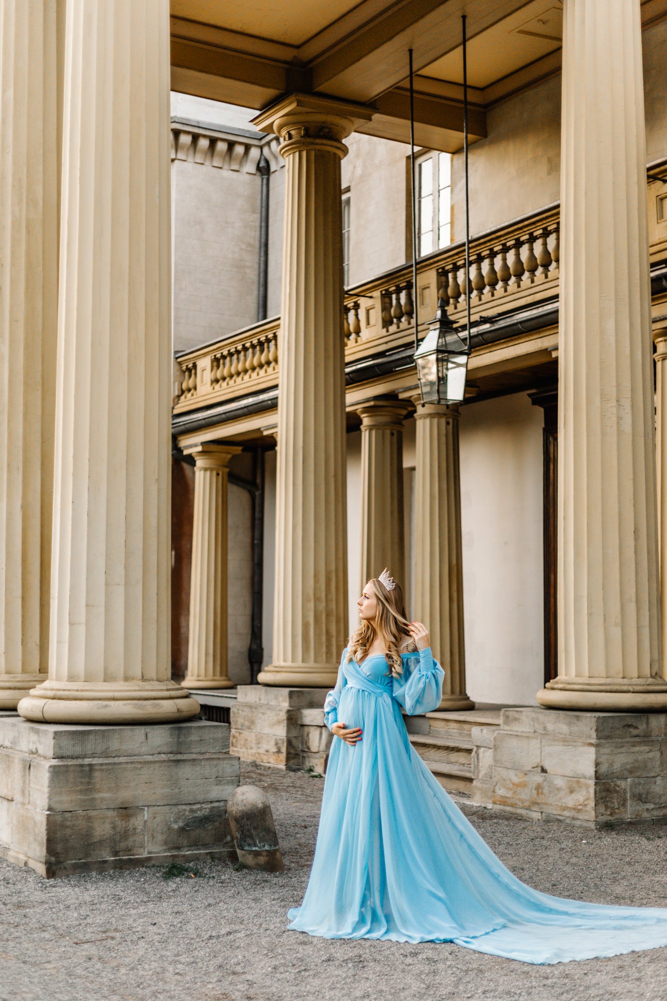 A woman in a flowing blue gown stands under tall classical columns. She is wearing a tiara and appears to be pregnant, holding her belly with one hand.