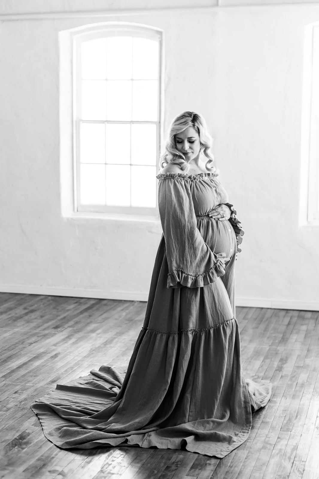 Black and white photo of a pregnant woman in a long dress, posing indoors with a wooden floor and large windows in the background.