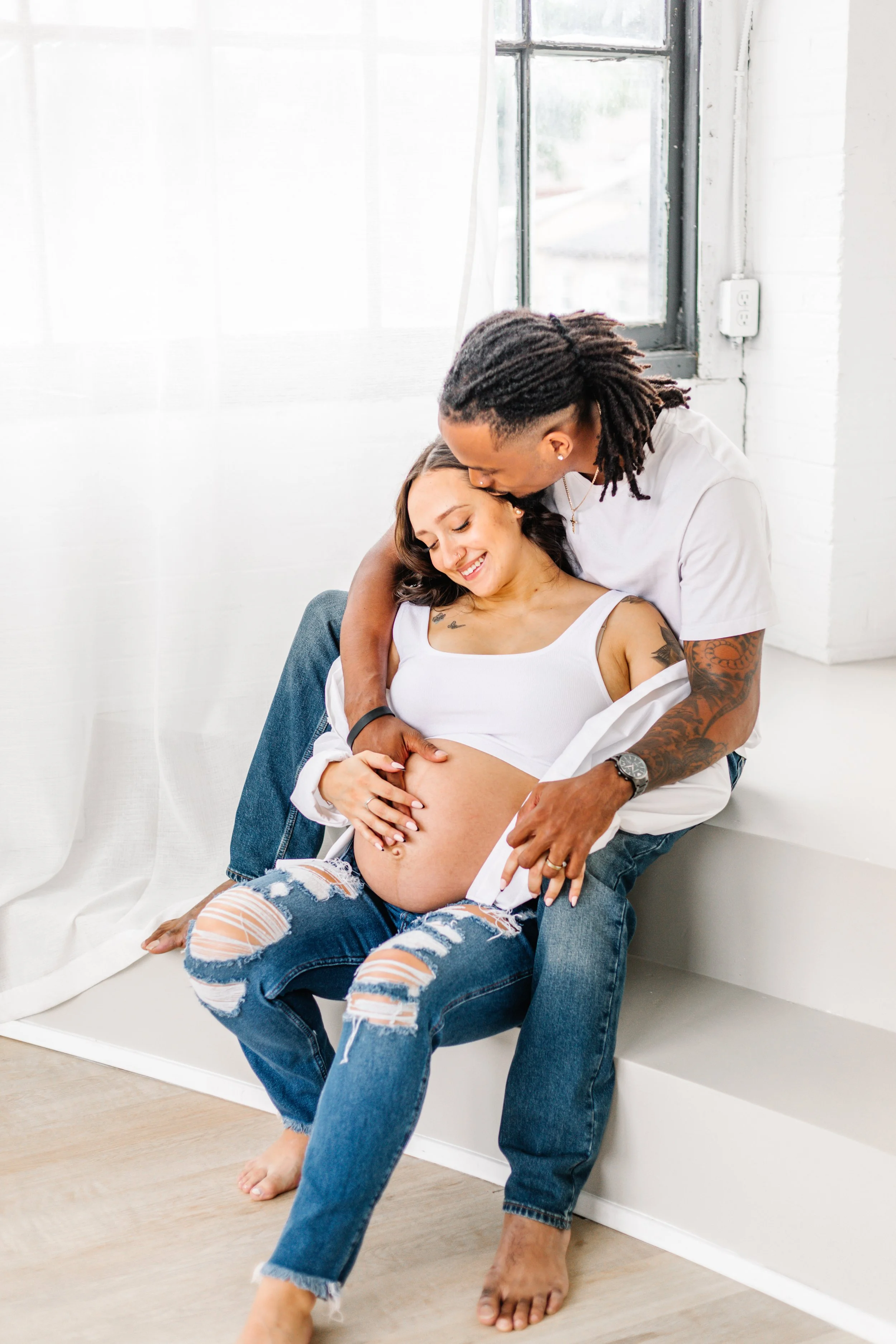 Pregnant woman sitting on steps with partner embracing her, both smiling, in casual attire.