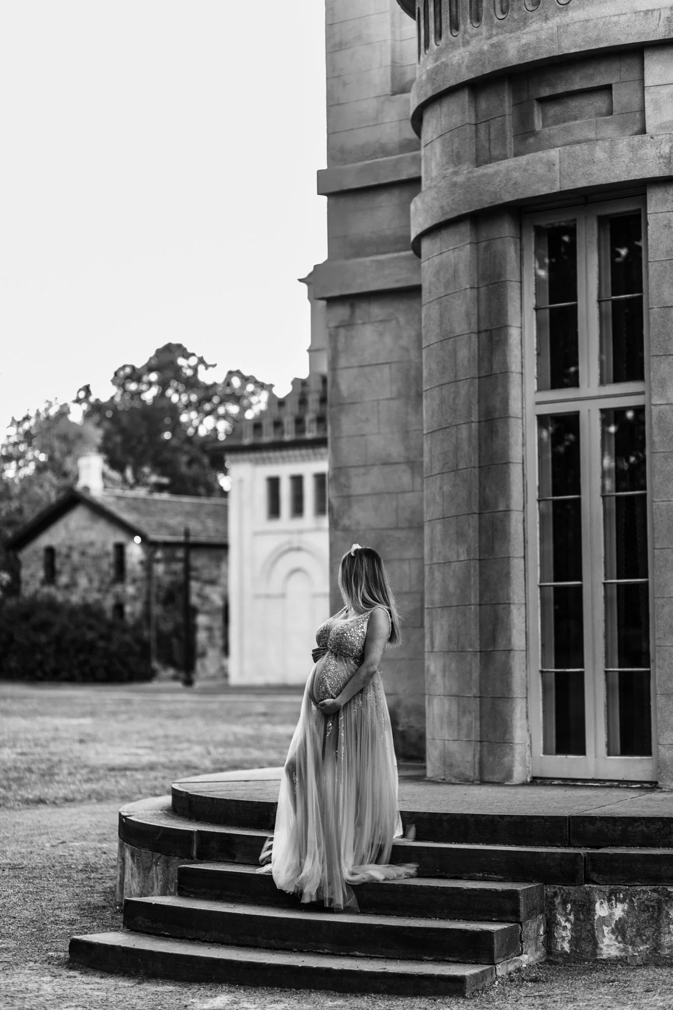 Pregnant woman in a flowing dress standing on steps next to a stone building in a black and white photo.