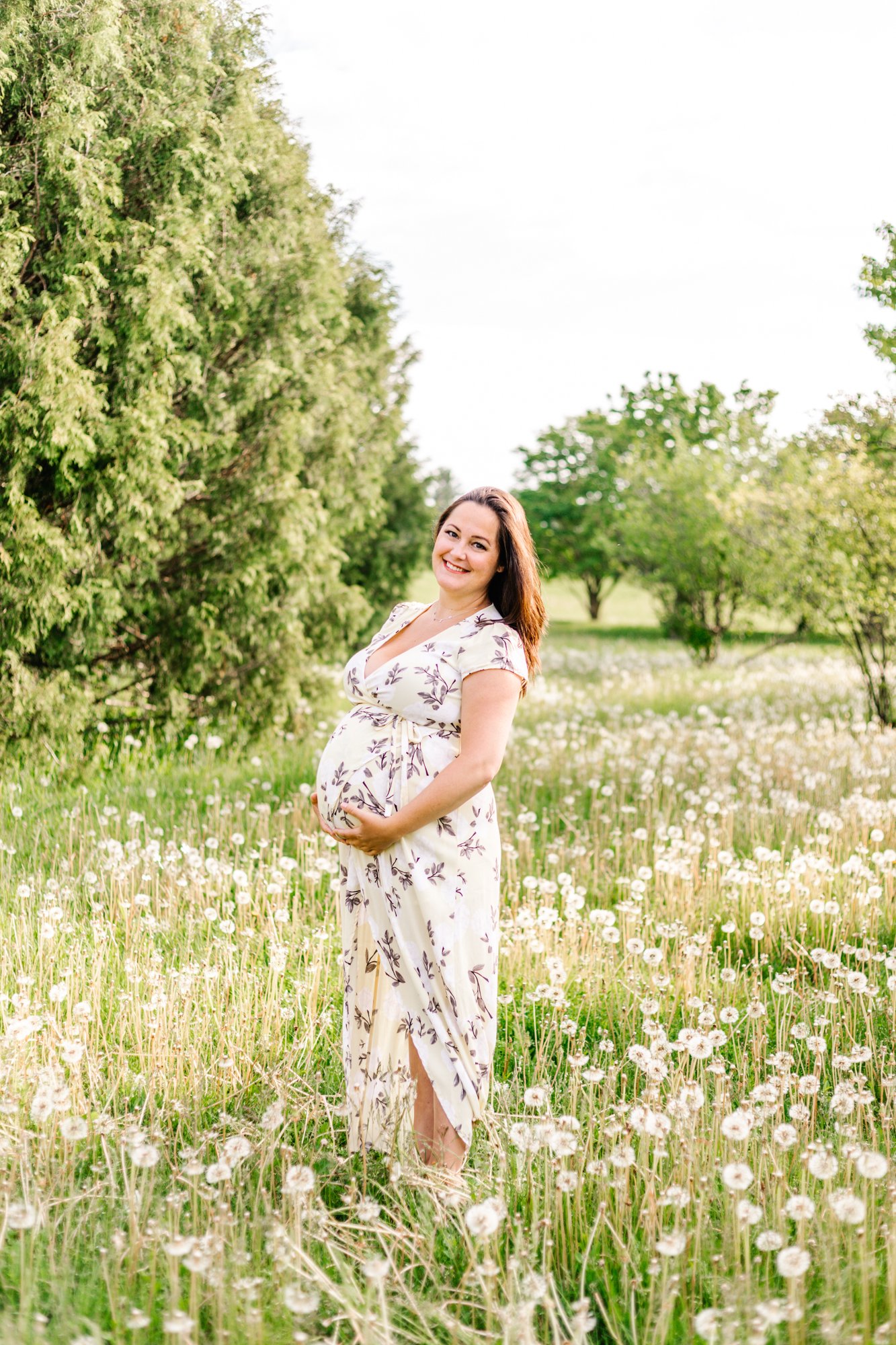 Pregnant woman in white floral dress standing in a field with dandelions and trees.
